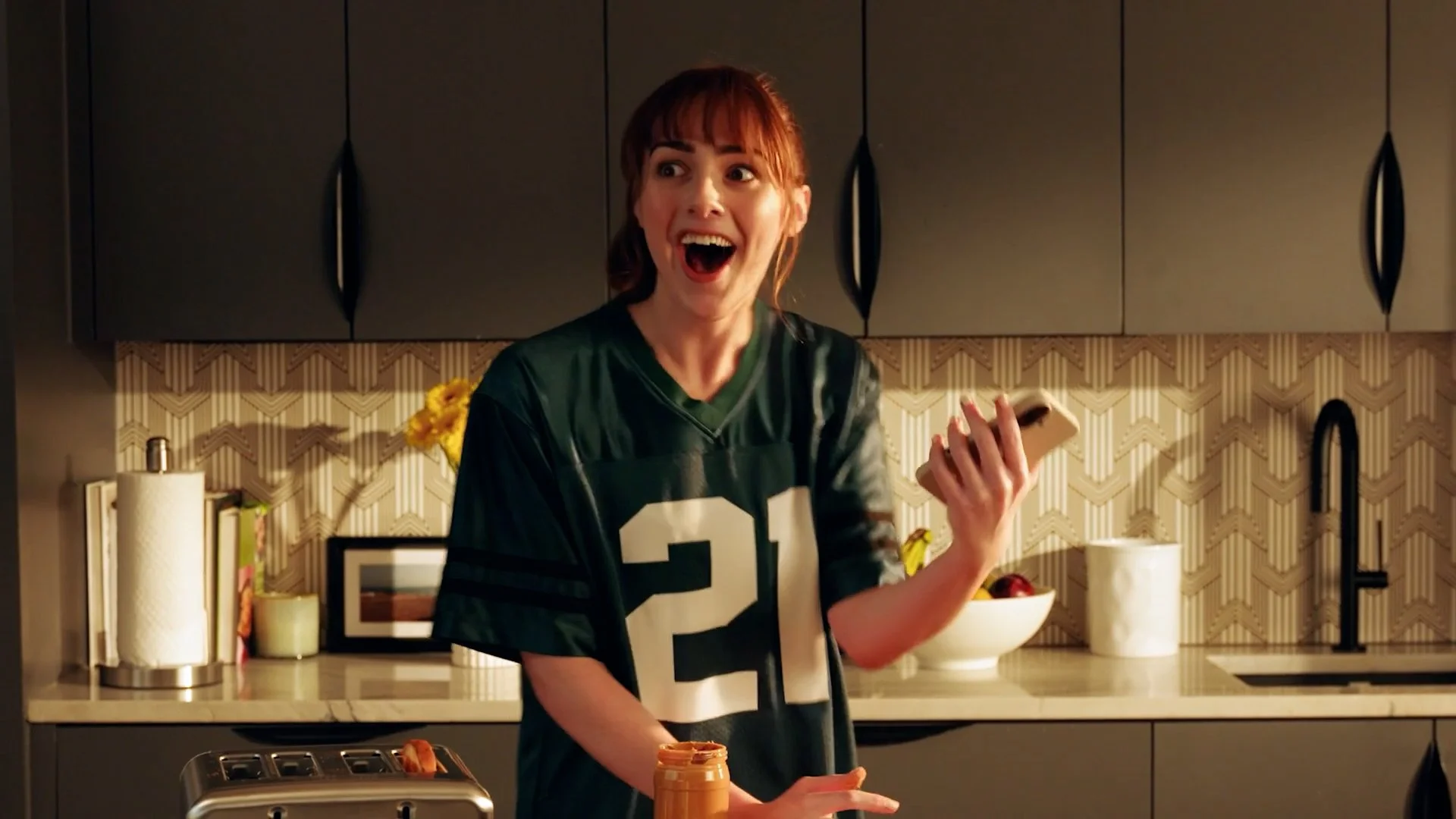 A woman with red hair wearing a green sports jersey with the number 21, smiling and holding a smartphone in her right hand while standing in a modern kitchen.