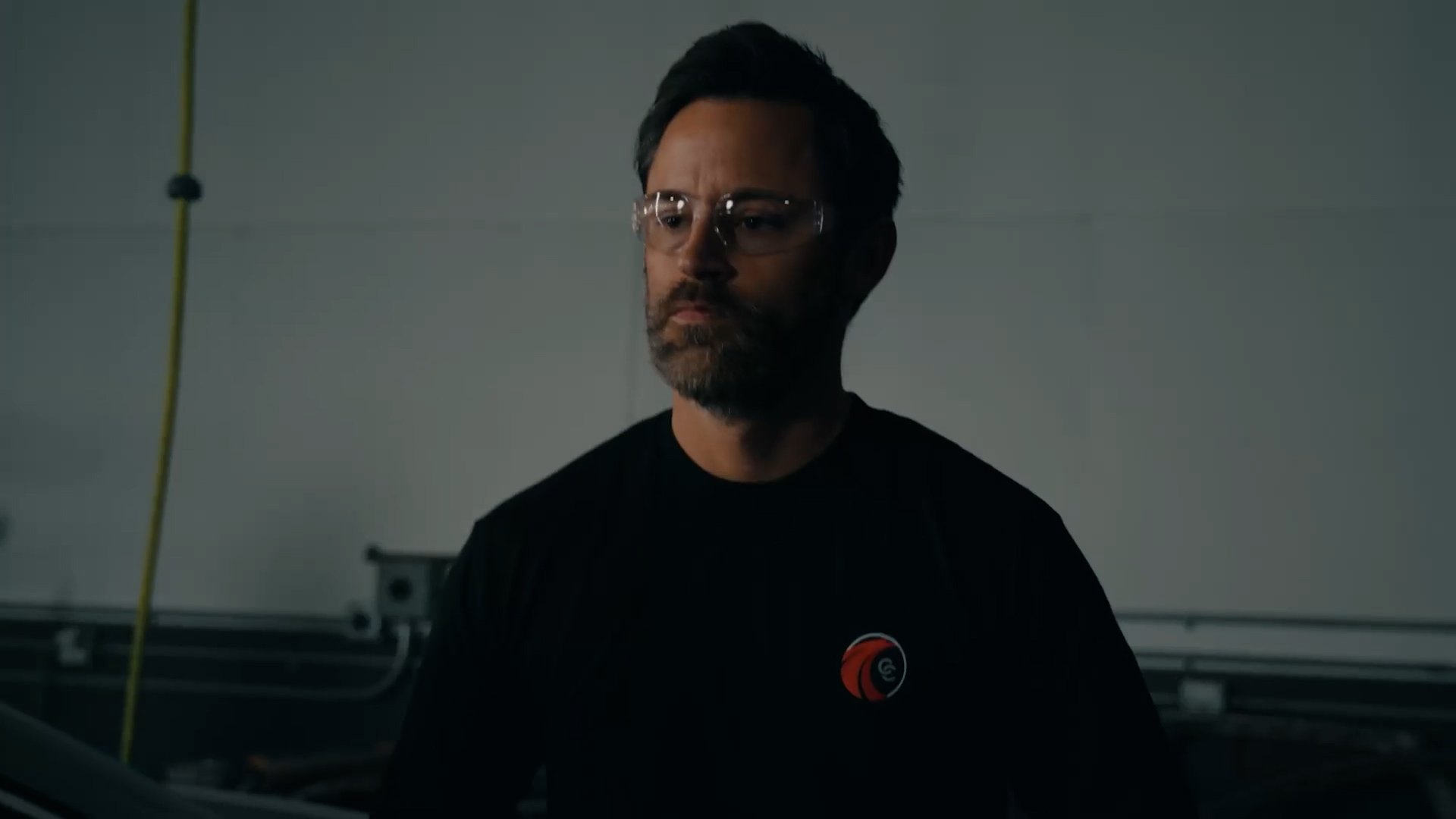 A man with dark hair, glasses, and a beard, wearing a black shirt with a red and black logo, standing in a dimly lit industrial or warehouse setting.