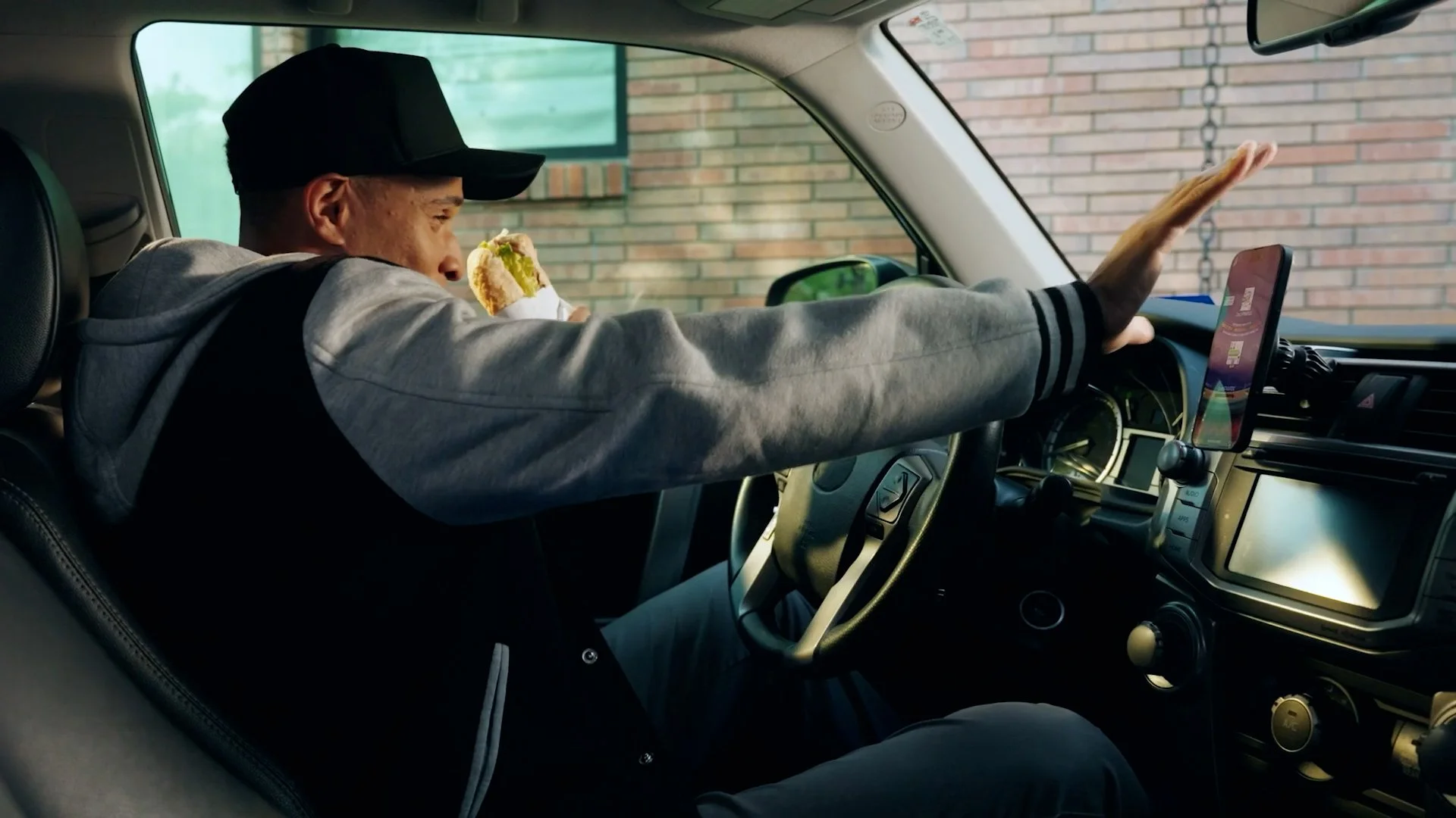 Man in a baseball cap and gray hoodie sitting in the driver's seat of a vehicle, holding a sandwich in his right hand and a phone mounted on the dashboard with his left hand, with a brick wall outside the vehicle.