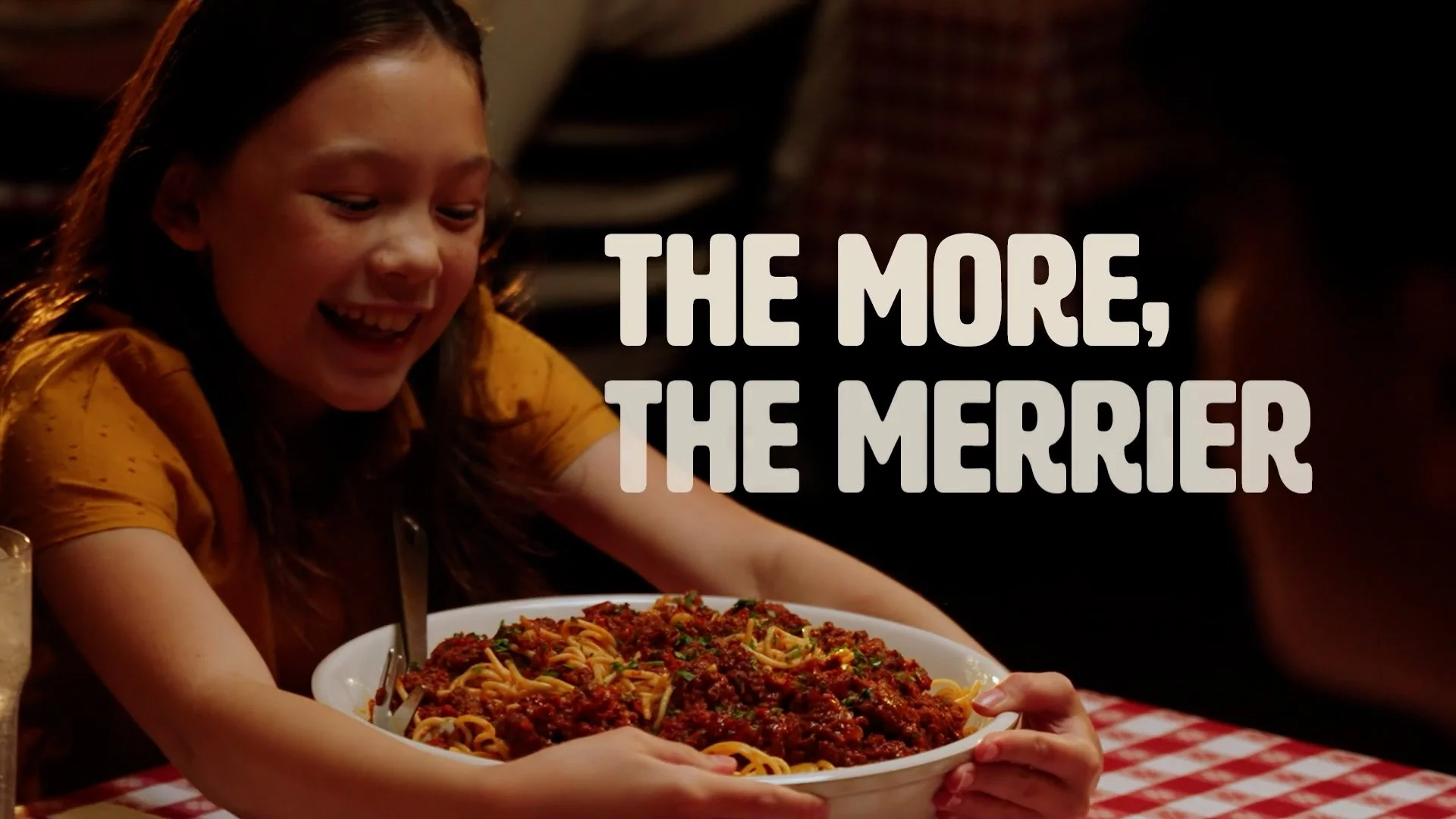 A young girl with brown hair smiling excitedly while holding a large plate of spaghetti with meat sauce and herbs, sitting at a table covered with a red and white checkered tablecloth, in a restaurant setting.