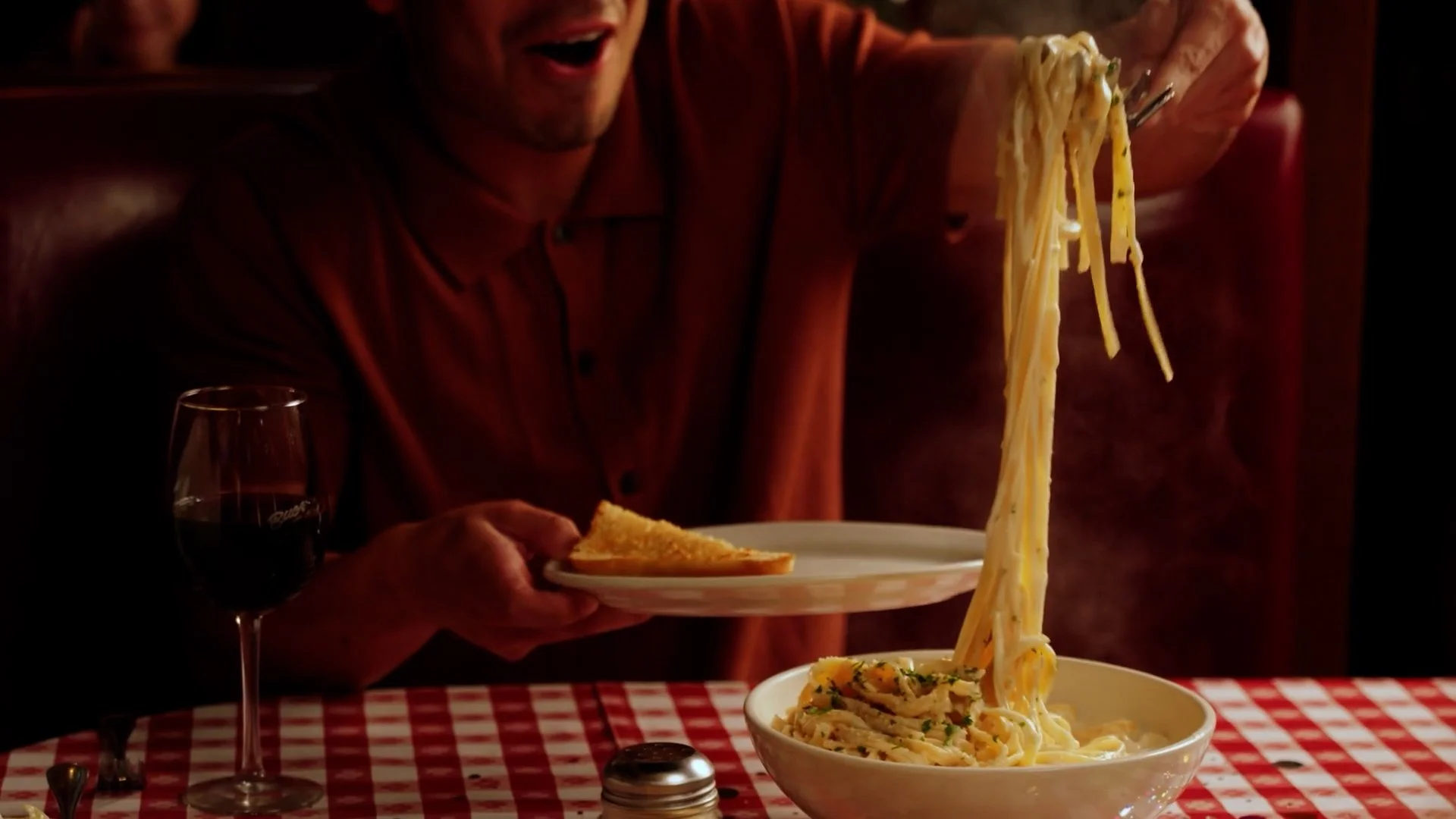 Person enjoying a bowl of spaghetti with grated cheese, at a table with a glass of red wine and a slice of garlic bread, in a cozy restaurant setting with a red checkered tablecloth.