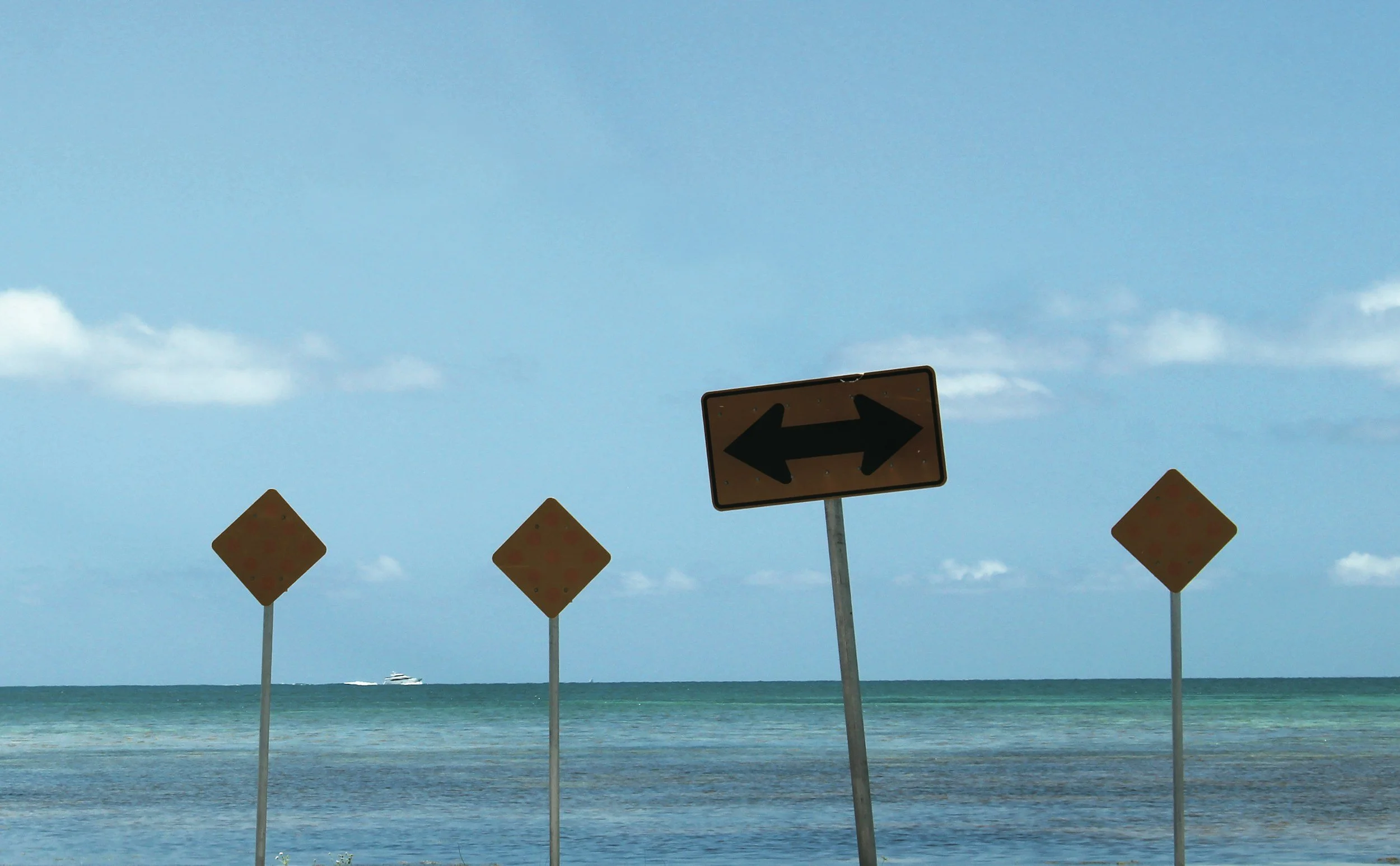 A beach scene with multiple road signs, including a large directional sign with black arrows pointing left and right, and three smaller diamond-shaped signs on poles in front of the ocean with a boat visible in the distance under a partly cloudy sky.