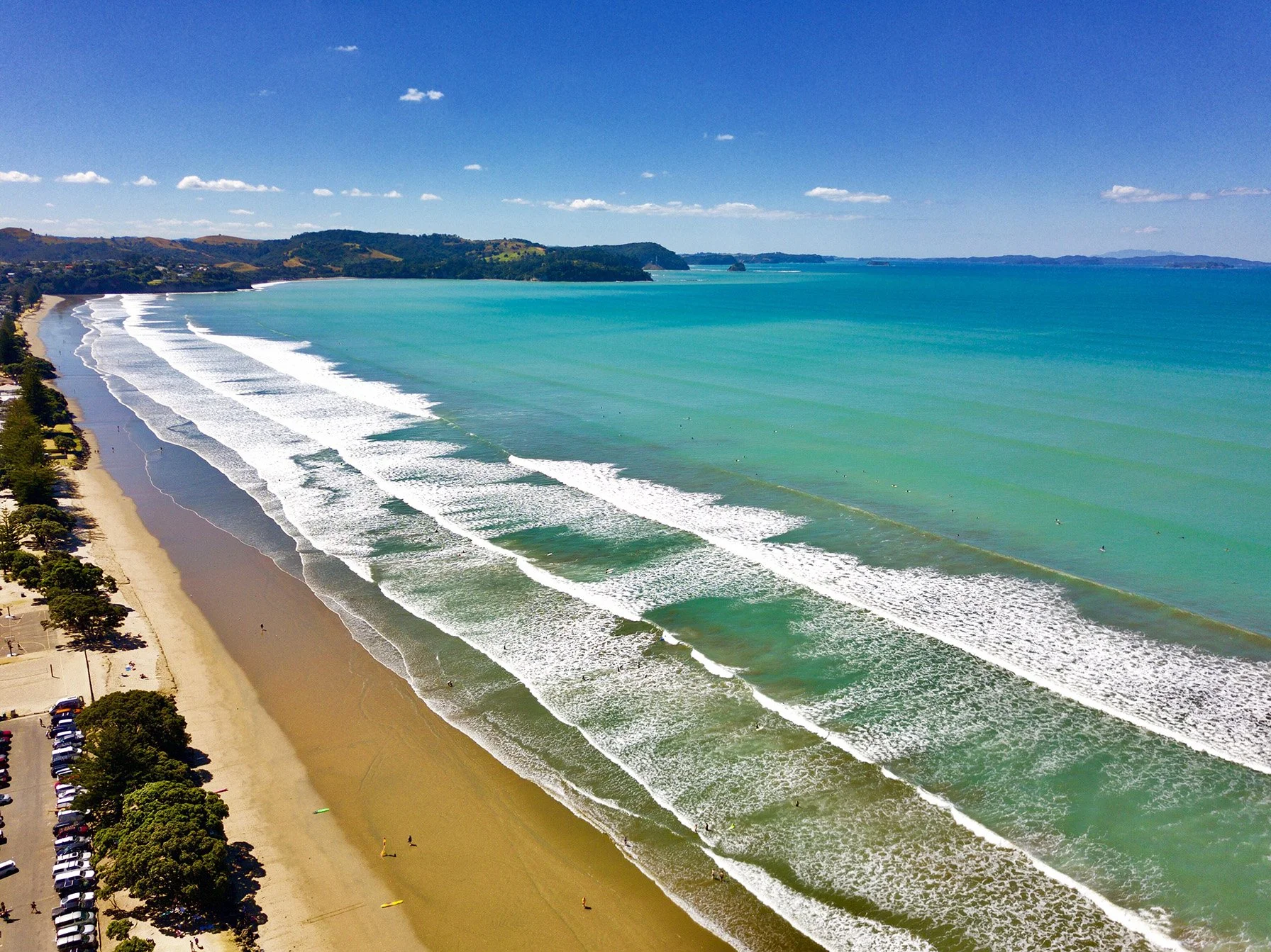 Aerial view of a sandy beach with green trees along the shore, adjacent parking lot, then the ocean with multiple surfable waves, under a clear blue sky with scattered clouds.