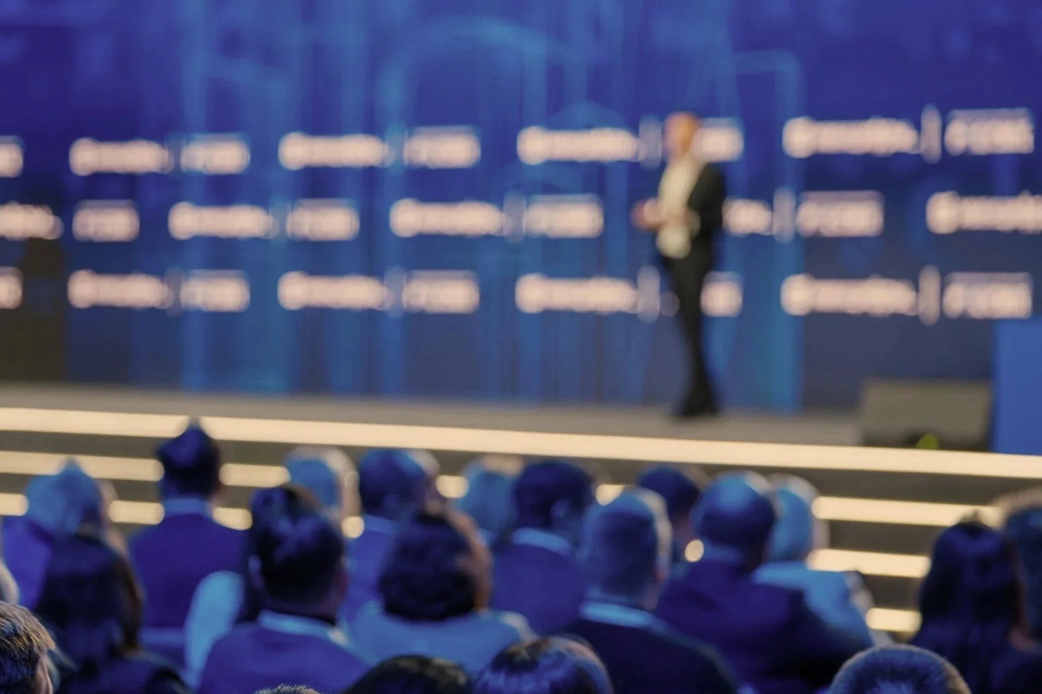 A speaker stands on stage delivering a presentation to a seated audience in a conference hall, with a large backdrop displaying a blue logo or text.