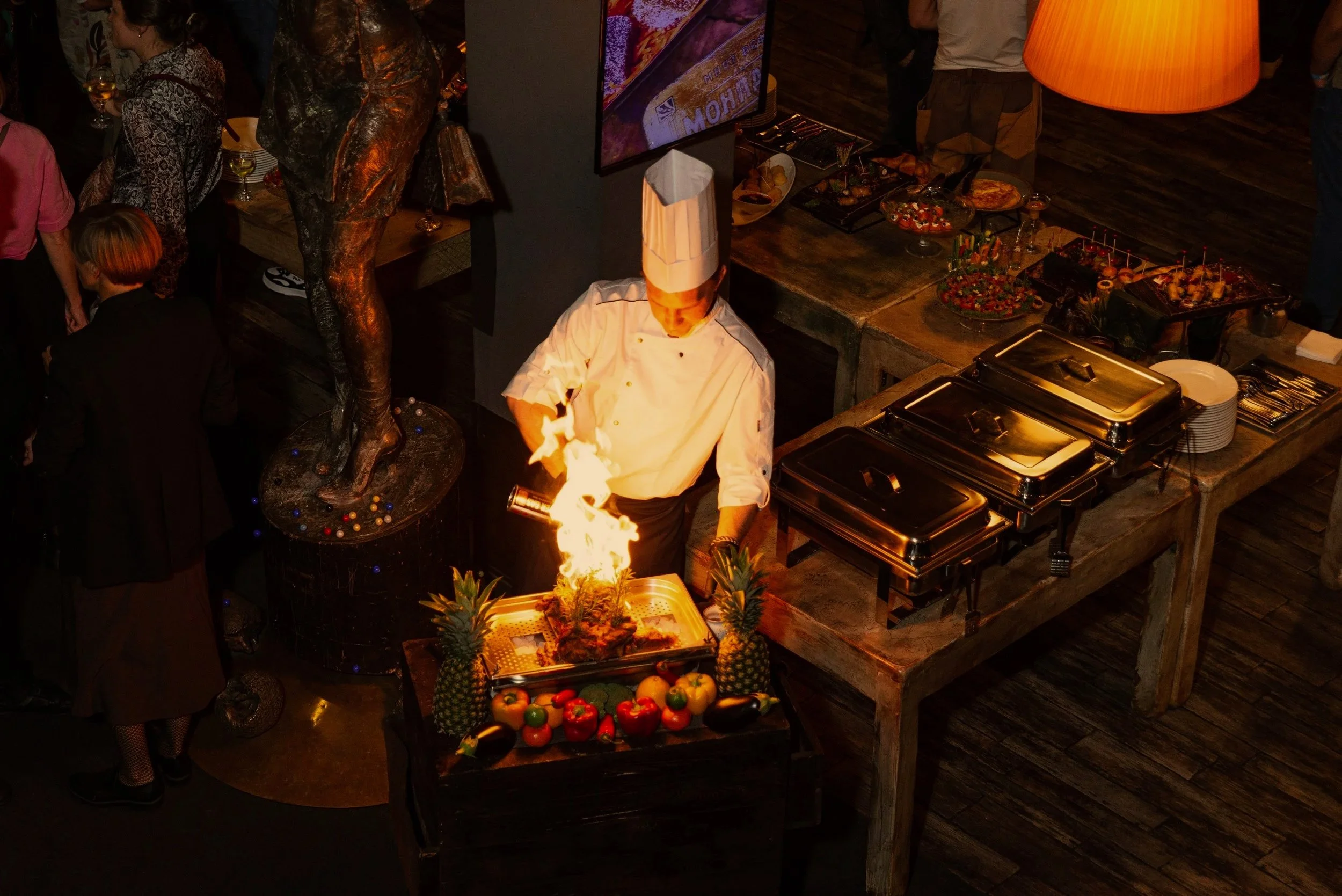 Chef in white uniform and tall hat cooking with flames at a buffet with tropical fruit decoration, including pineapples, peppers, and eggplants, in a restaurant setting with guests and food stations.