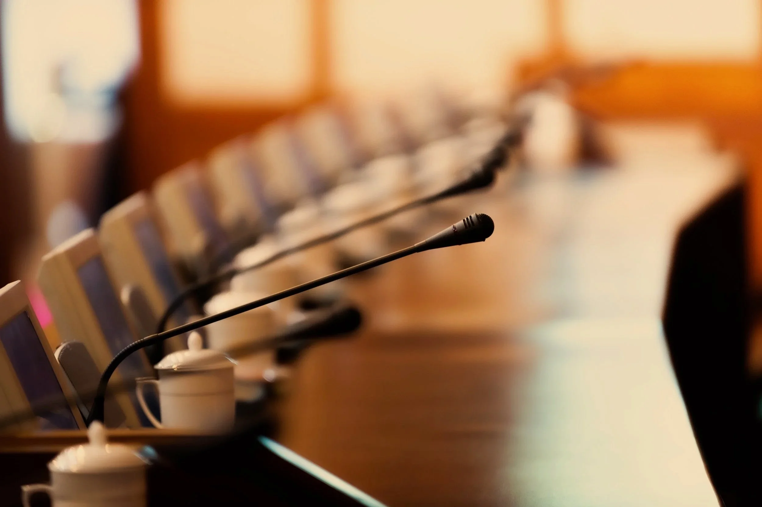 A row of microphones on a conference table with cups in front of each microphone, conference room with wooden paneling in the background.