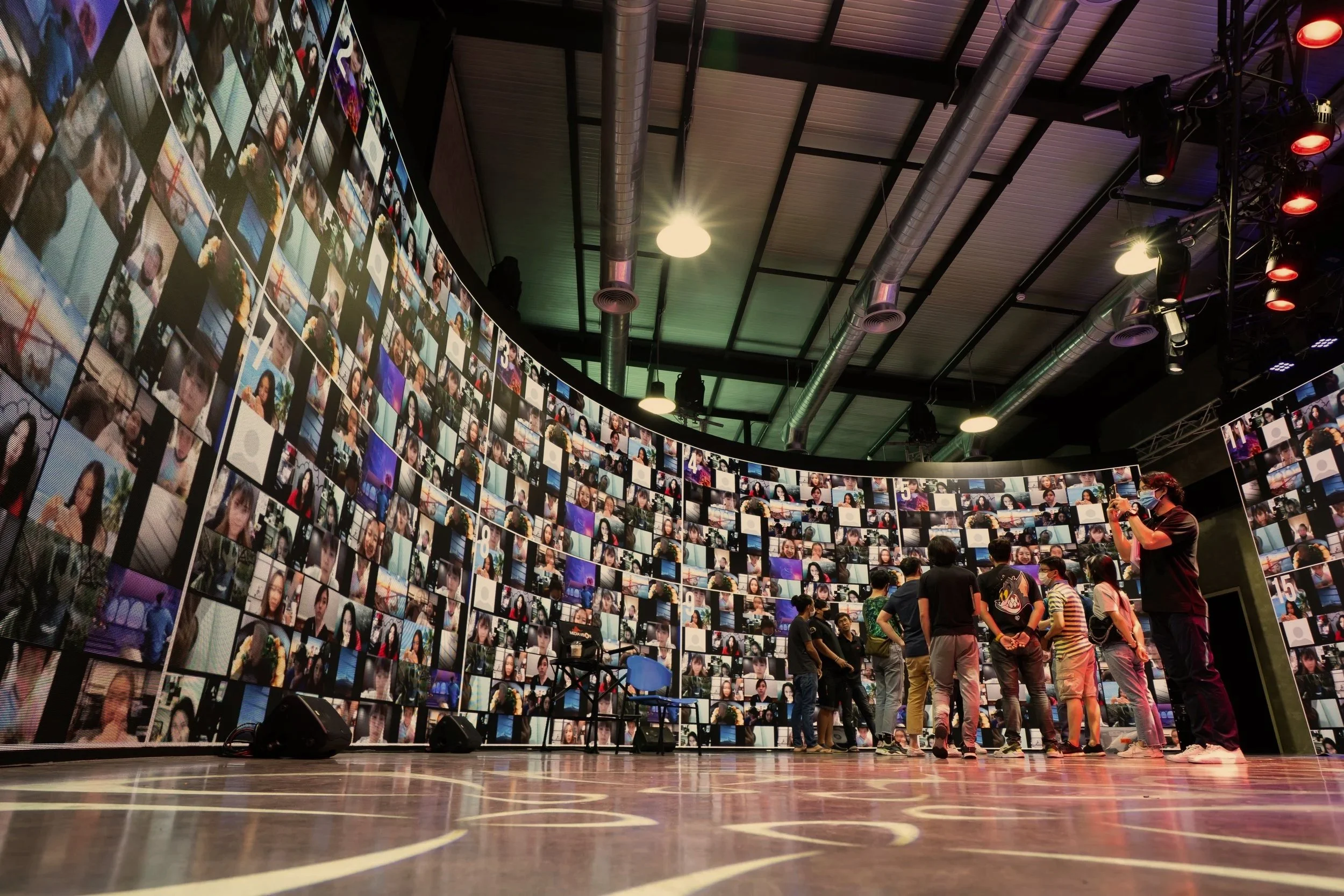 A large curved digital screen displaying numerous small individual photos of people surrounds a group of people standing on a stage. One person appears to be taking a picture, and there are chairs and speakers on the stage. The ceiling has exposed pipes and industrial-style lighting.