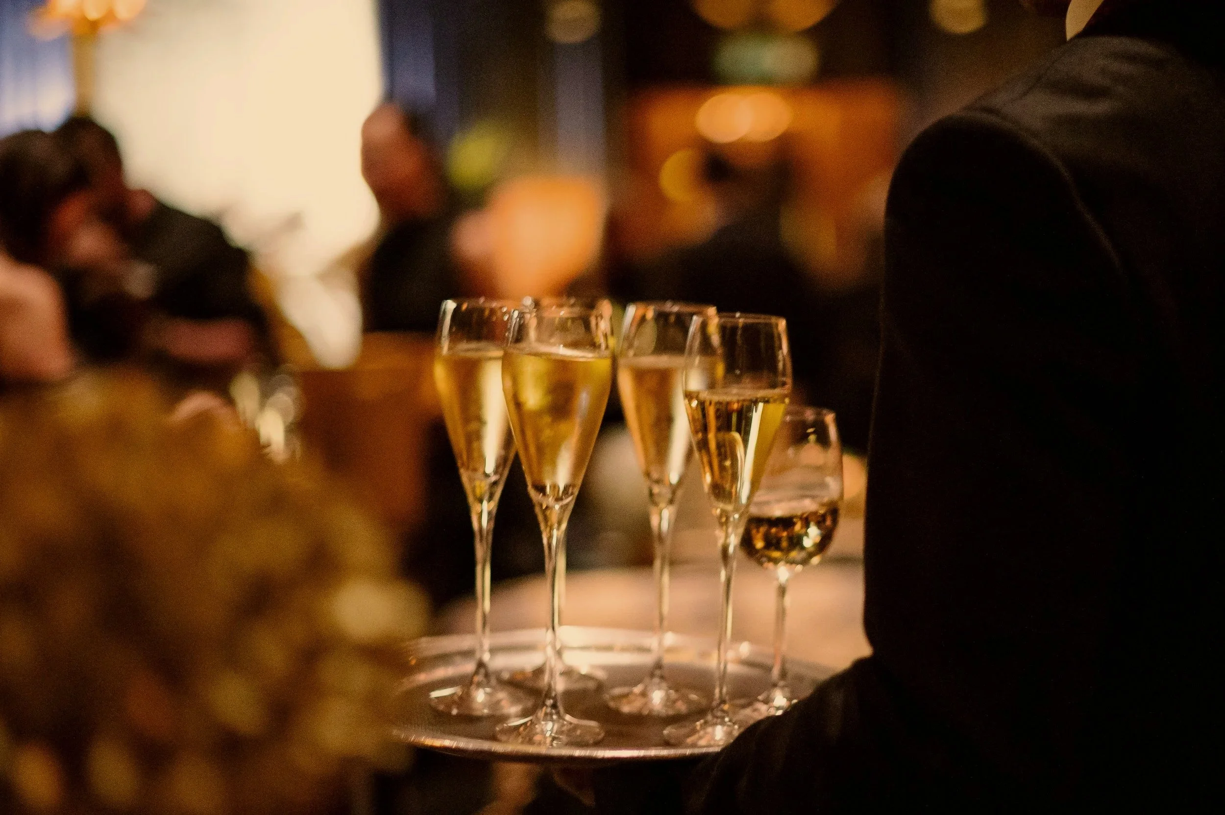 A server in a black suit holding a tray of five champagne glasses with sparkling wine, with other people blurred in the background at a social gathering or celebration.