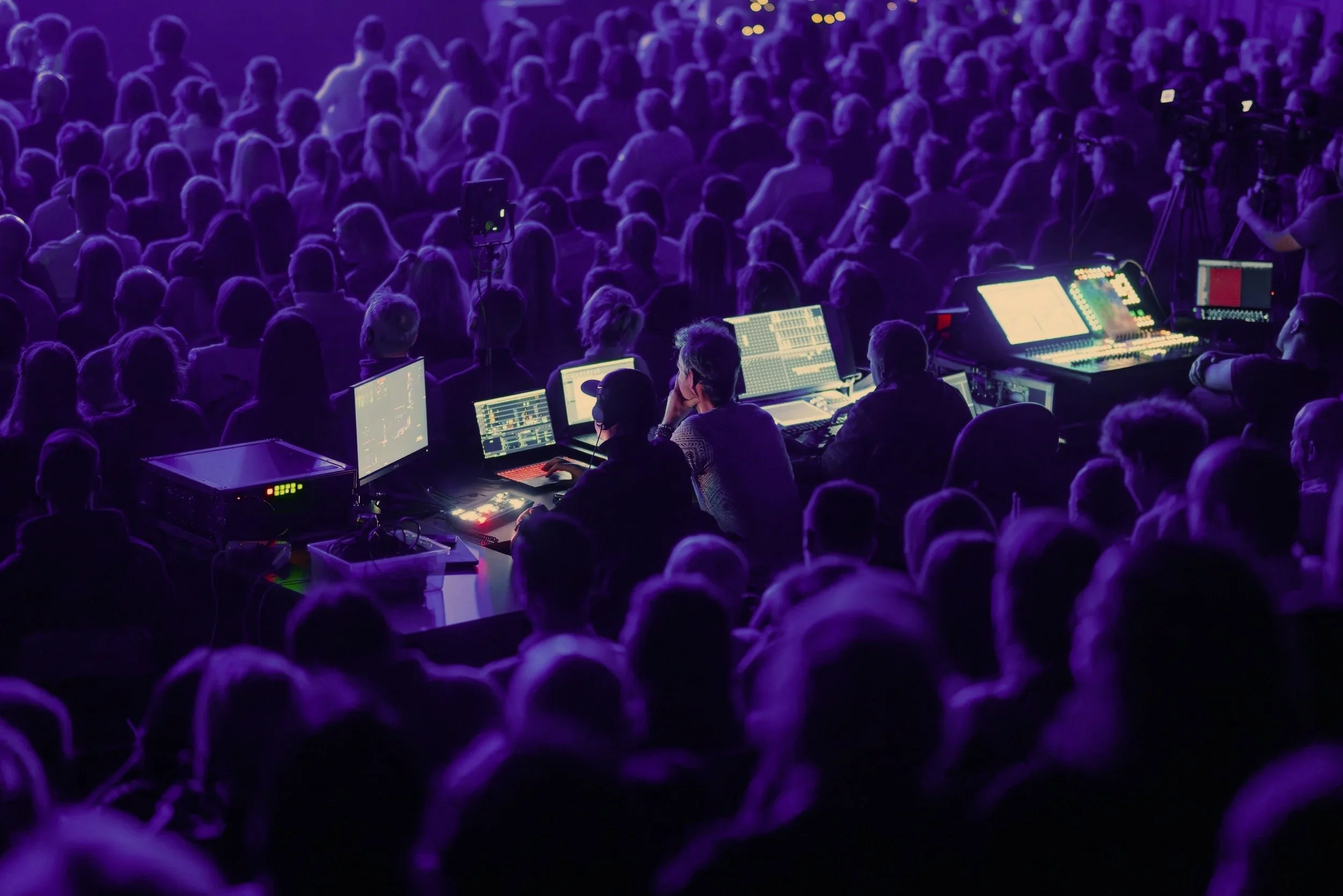 A large crowd of people attending a concert or event, with a group of sound and lighting technicians operating equipment in the foreground, illuminated by purple lighting.