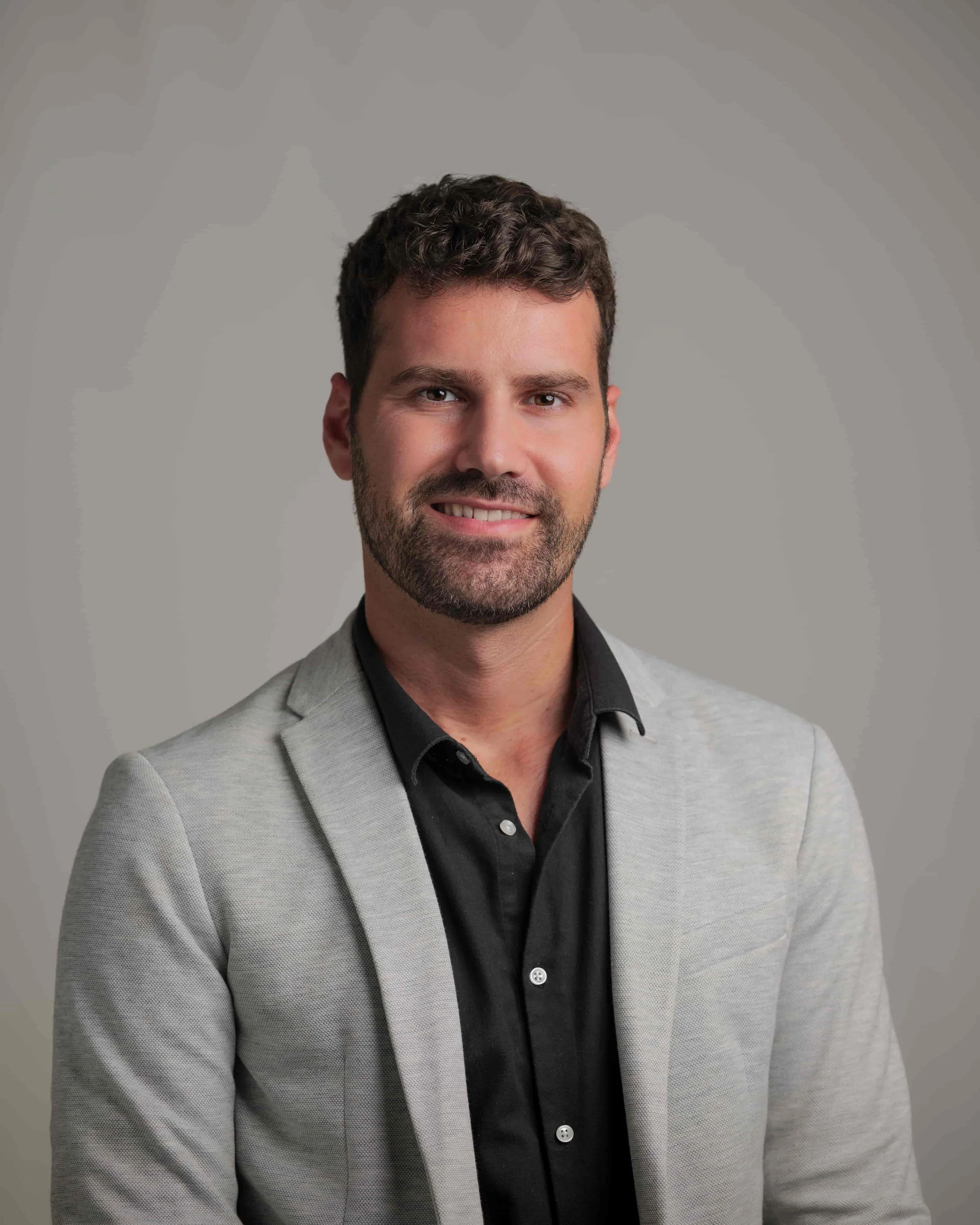 Man wearing a beard wearing a blazer  and a black shirt, smilling with a grey background.