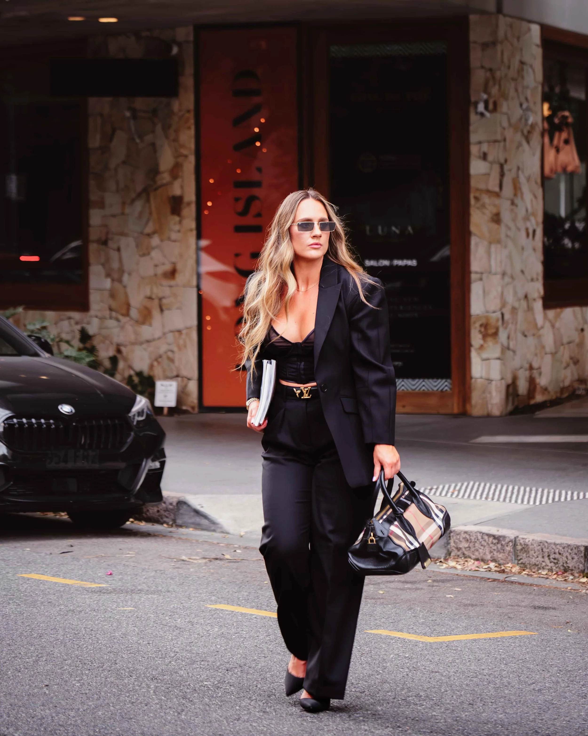 A sophisticated woman in a tailored black power suit and dark lenses, carrying a chic handbag and a book as she navigates the city streets.