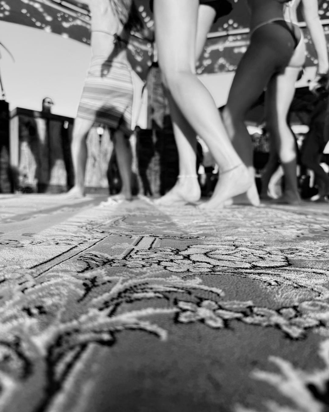 People walking barefoot on a decorated outdoor rug or mat, with some wearing swimsuits, possibly at a poolside or beach gathering.