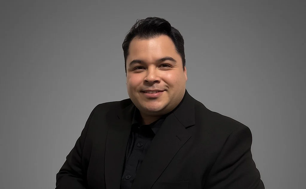 A man in a black suit smiling against a gray background.
