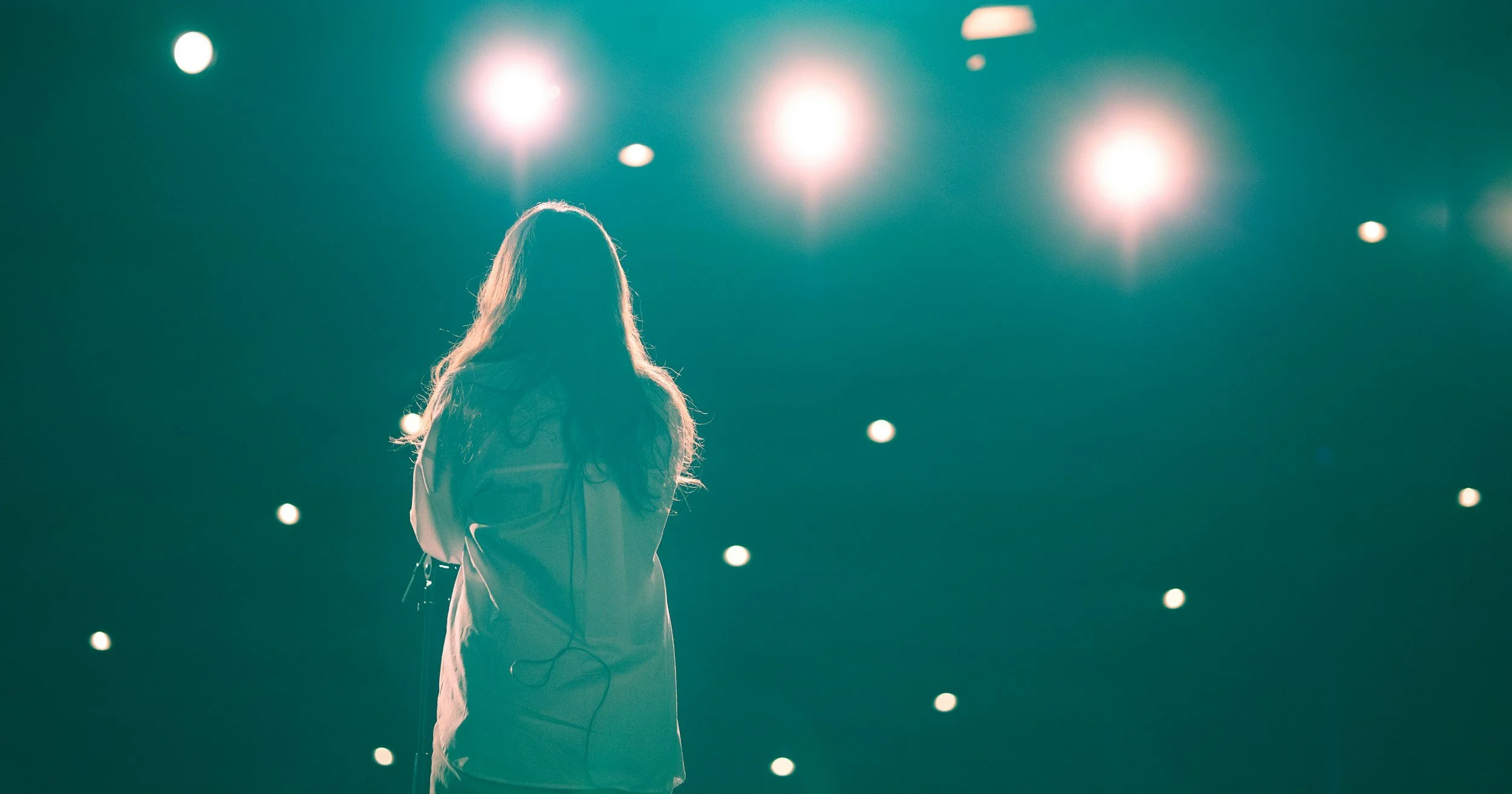 A person standing on a stage with bright lights overhead, seen from behind with long hair and a backpack.