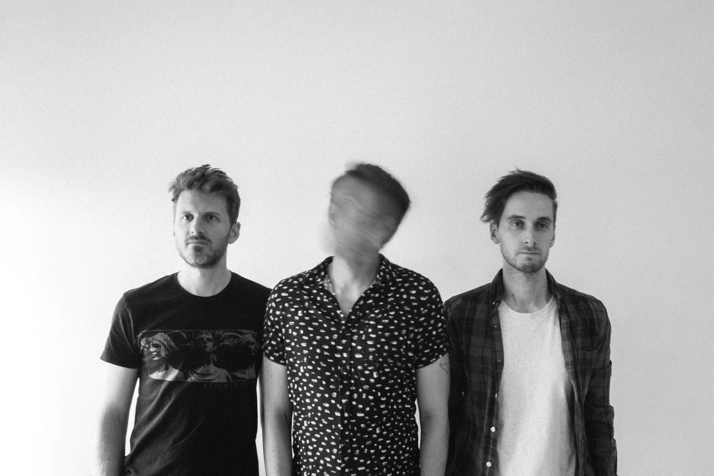Black and white photo of three men standing in front of a plain wall. The man on the left is wearing a t-shirt with a graphic, looks serious, has short hair and a beard. The man in the middle has a blurred face, is wearing a patterned shirt, and has short hair. The man on the right is wearing a casual shirt over a t-shirt, has longer hair, and a serious expression.