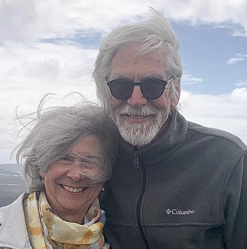 A smiling elderly couple outdoors on a cloudy day, with the man wearing sunglasses and a Columbia jacket, and the woman wearing a scarf and white jacket.