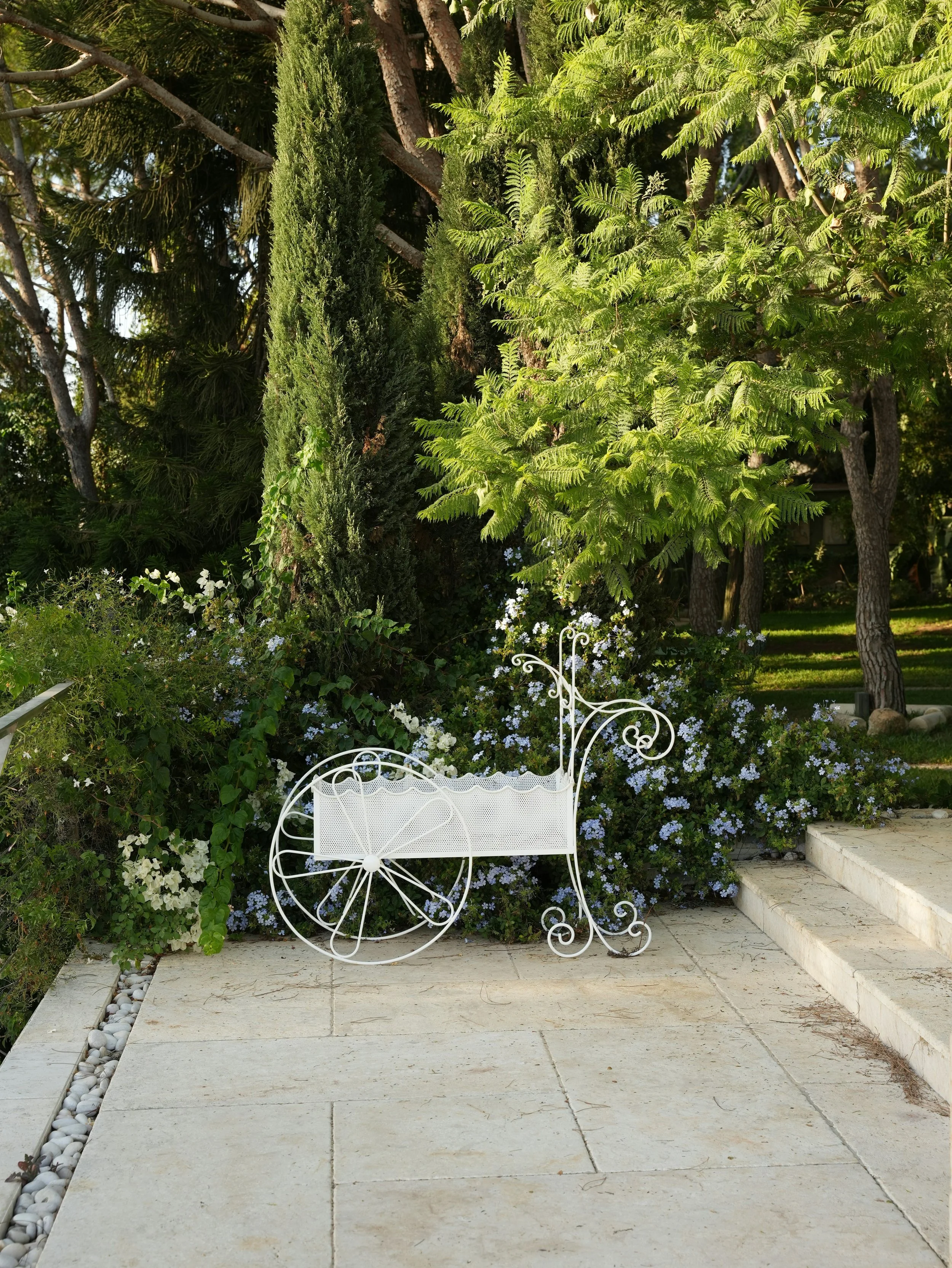 White decorative wrought iron garden bench shaped like a vintage baby carriage, situated on stone patio amidst lush green trees and flowering bushes.