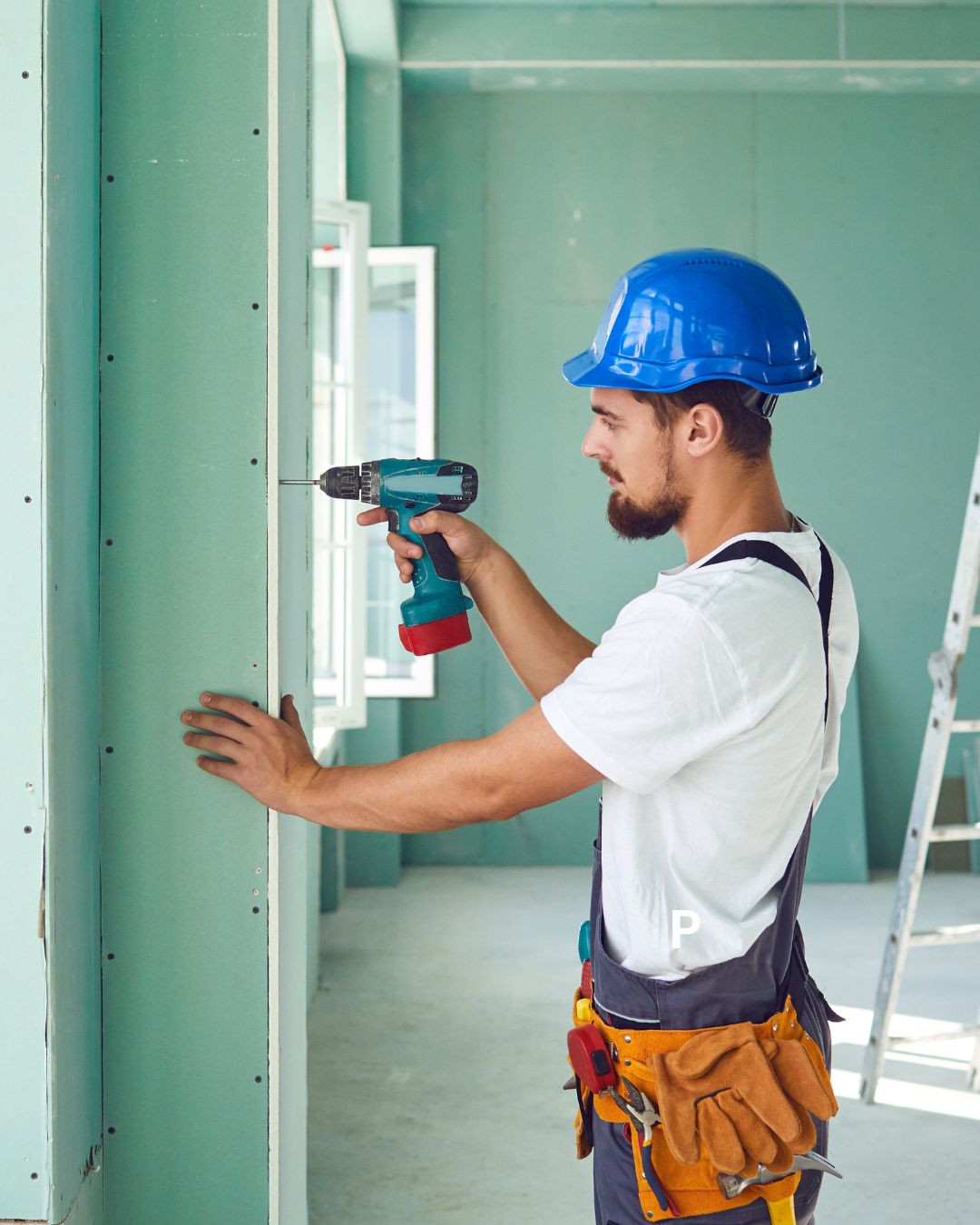 A construction worker with a blue hard hat using a cordless drill to install or secure panels in a building under construction.