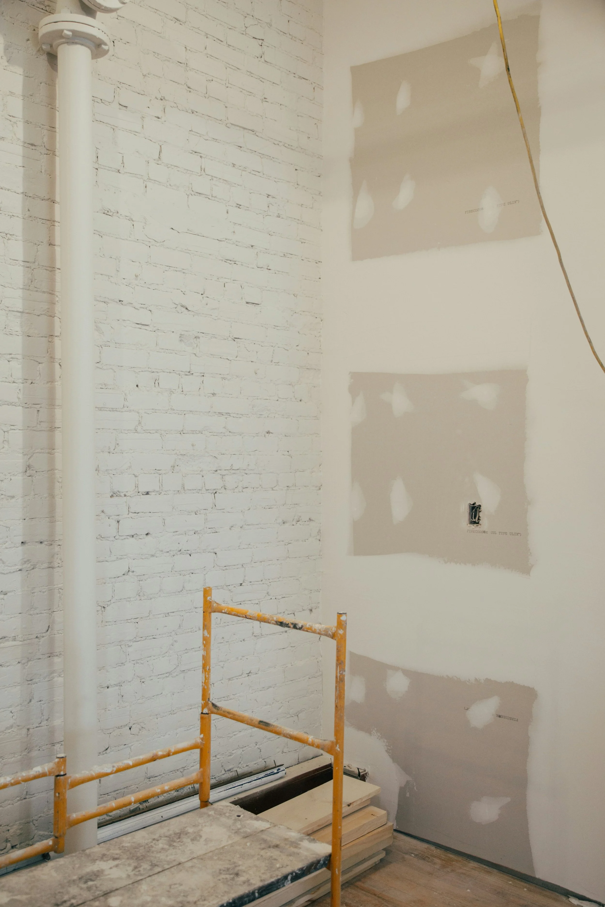 Interior of a room under construction with a white brick wall on the left, drywall with taped and mudded seams on the right, a yellow safety barrier, some wood planks, and a yellow electrical wire hanging from the ceiling.