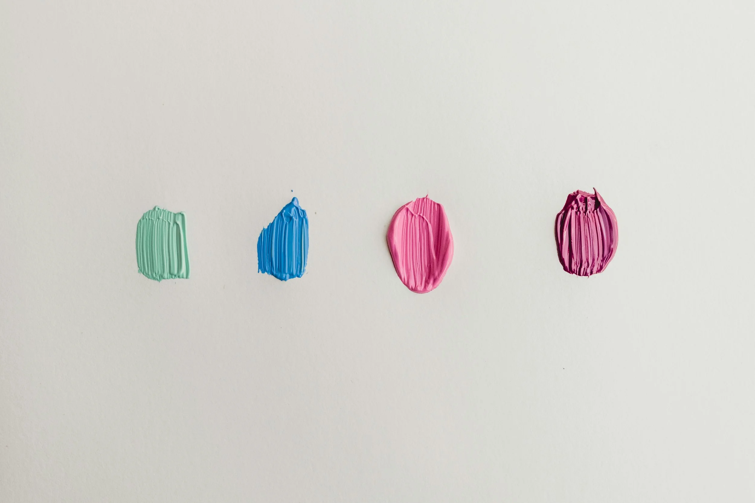 Four small dollops of paint in green, blue, pink, and purple on a white background.