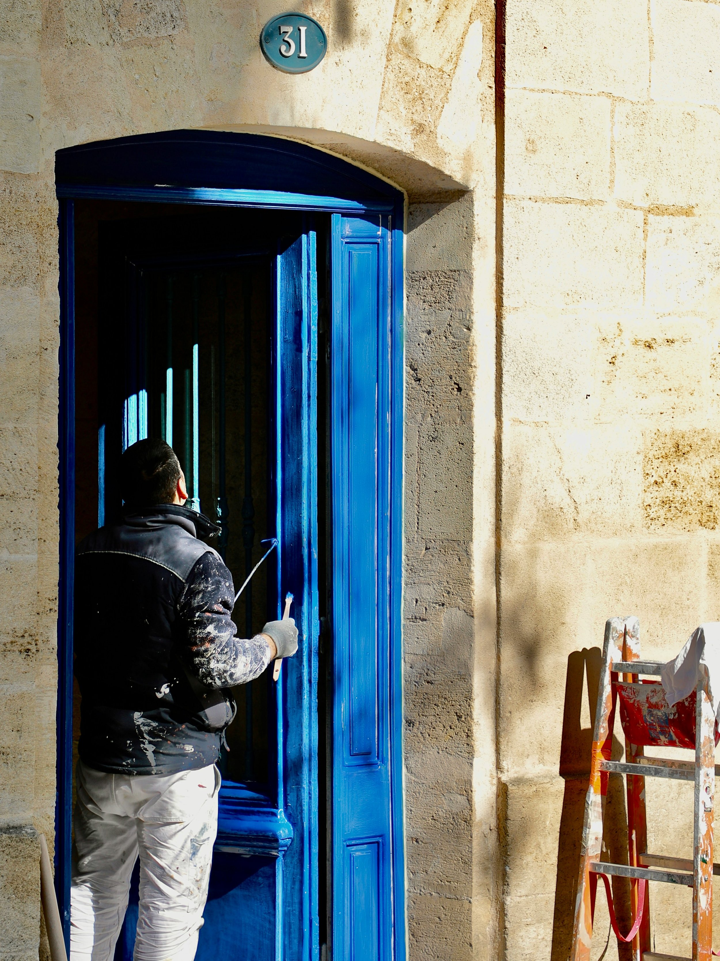 A man painting a blue door on a stone building with the number 31 above it. A ladder with paint splatters stands nearby.