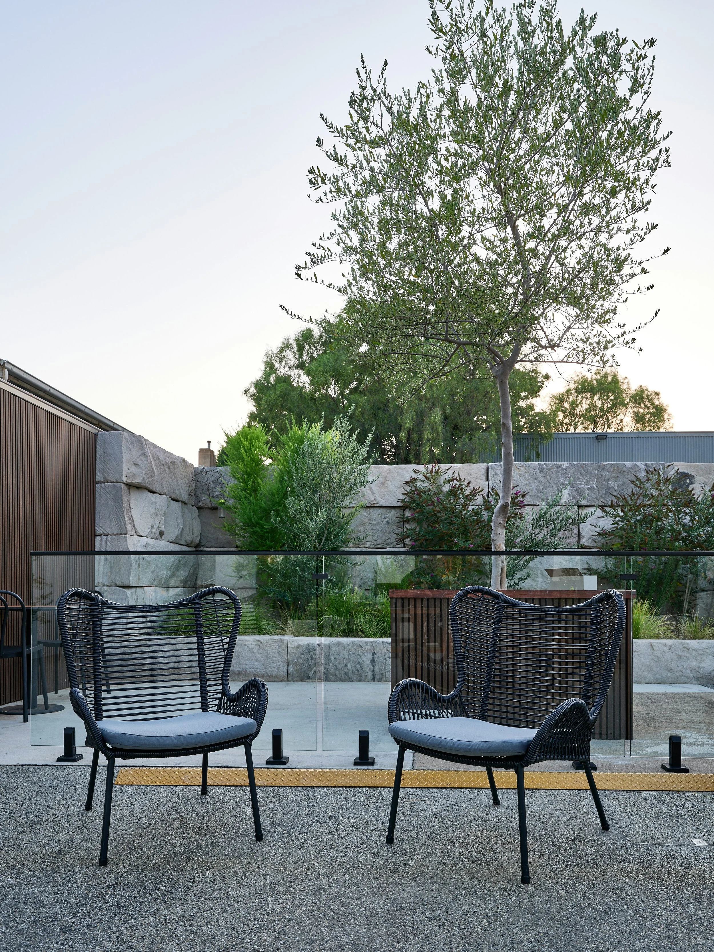 Two black outdoor chairs with gray cushions on a patio, overlooking a landscaped garden with trees, shrubs, and a stone wall, enclosed by glass panels.