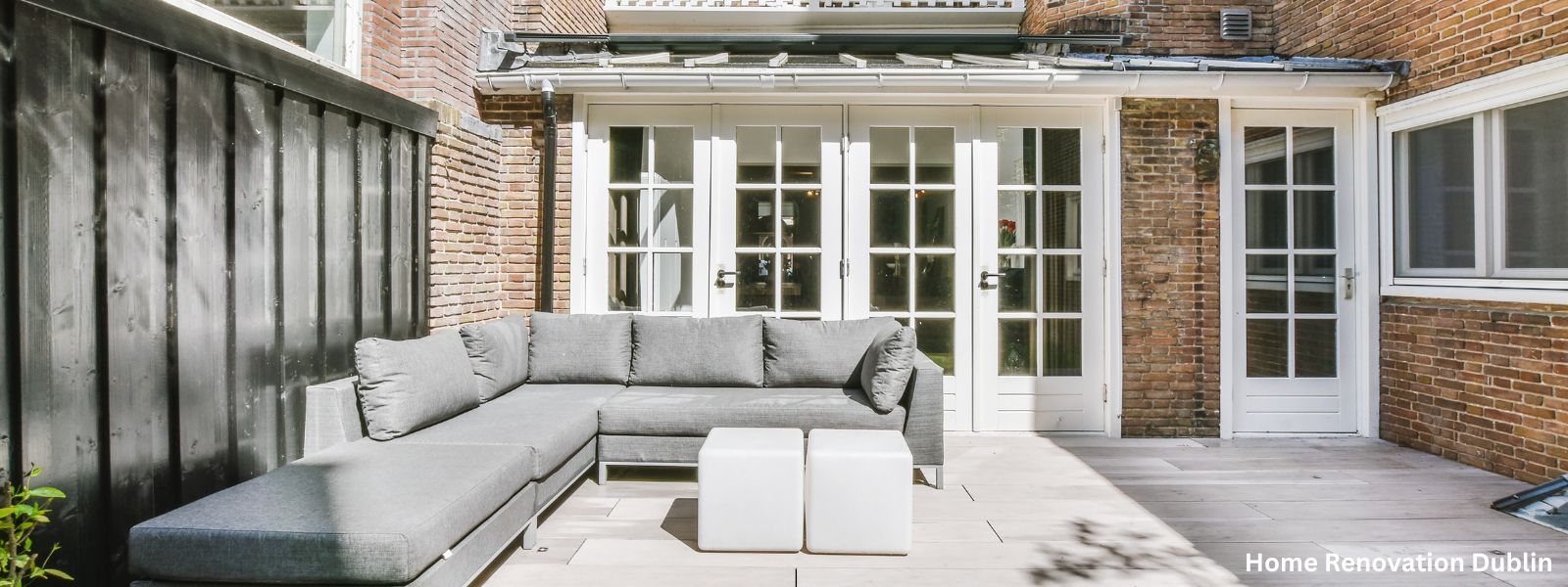 A modern outdoor patio with a gray sectional sofa, two white cube stools, and white French doors on a brick house with a wooden deck.