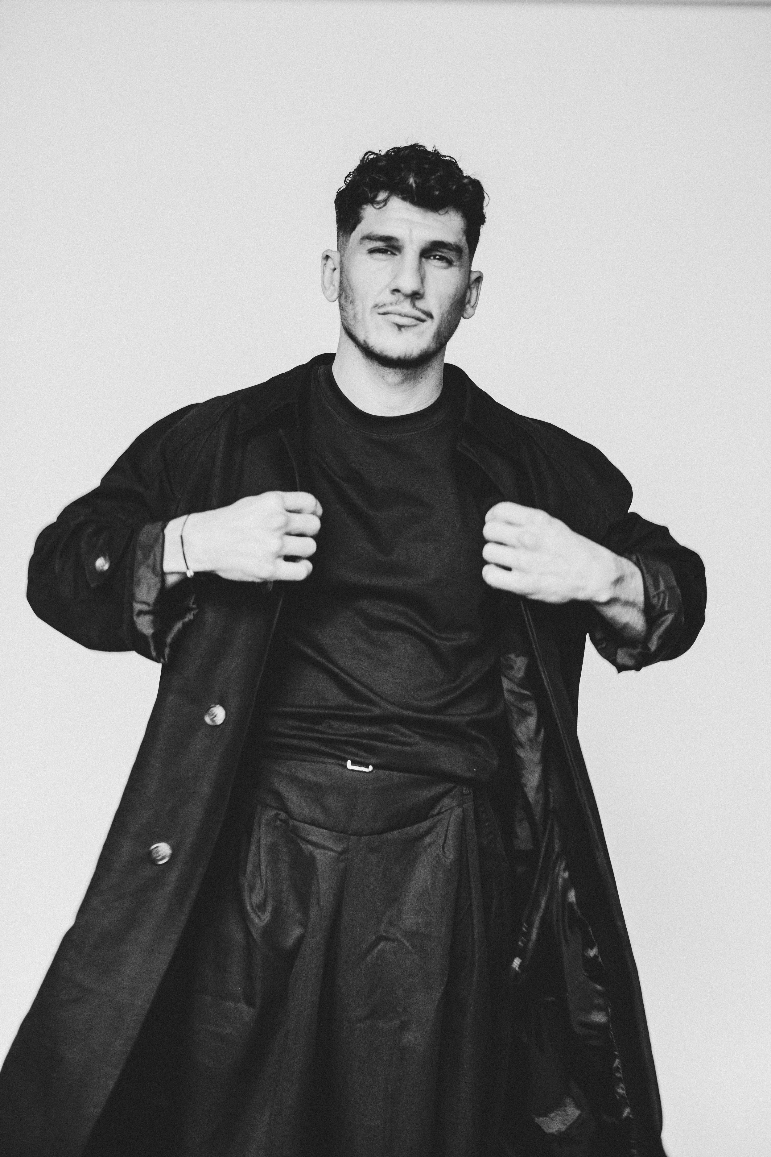 Un jeune homme en noir et blanc portant une veste ouverte, regardant vers l'objectif, contre un fond blanc uniform.