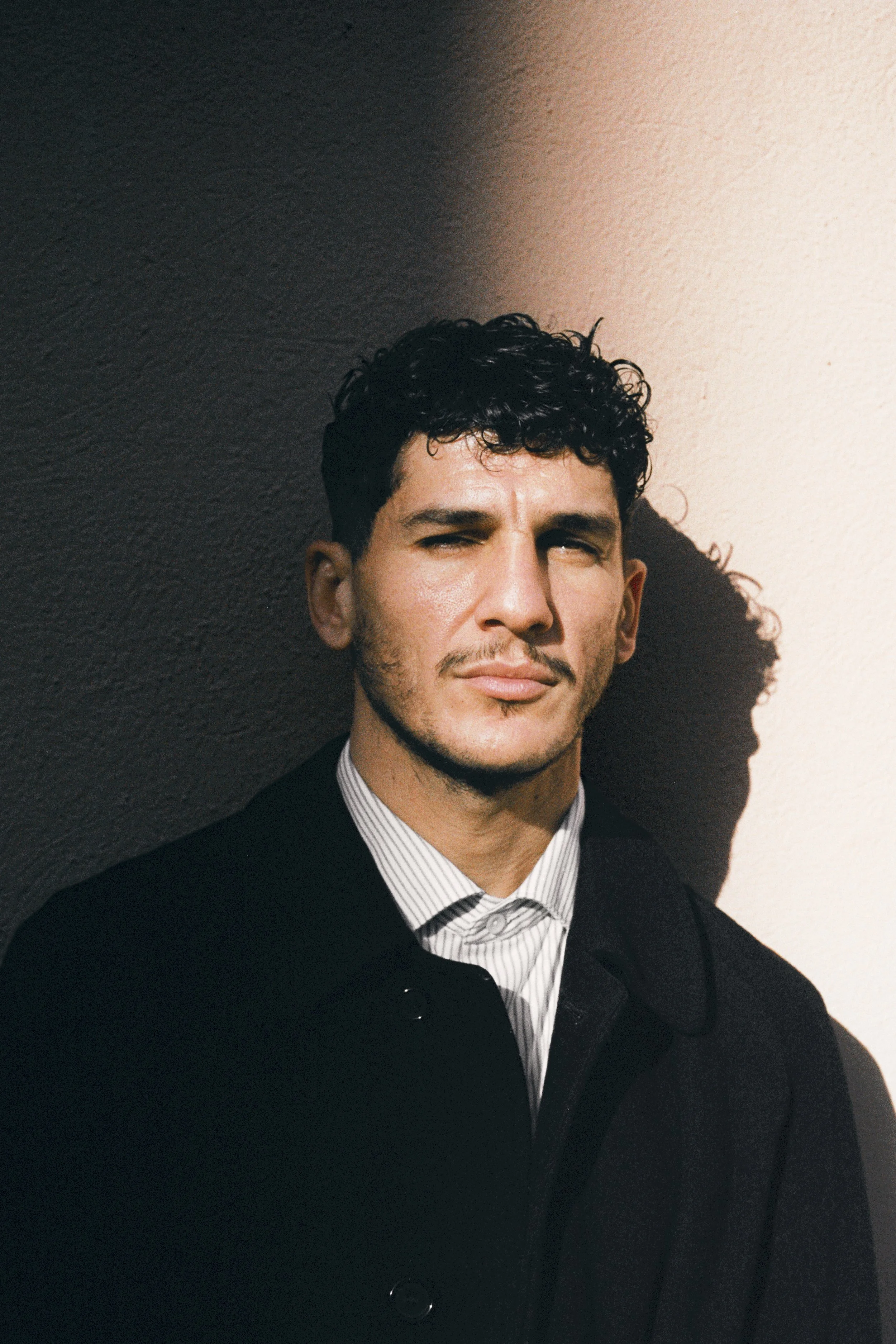 Un homme debout contre un mur, regardant légèrement de côté, avec un éclairage contrasté créant une ombre accompagnant sa silhouette.