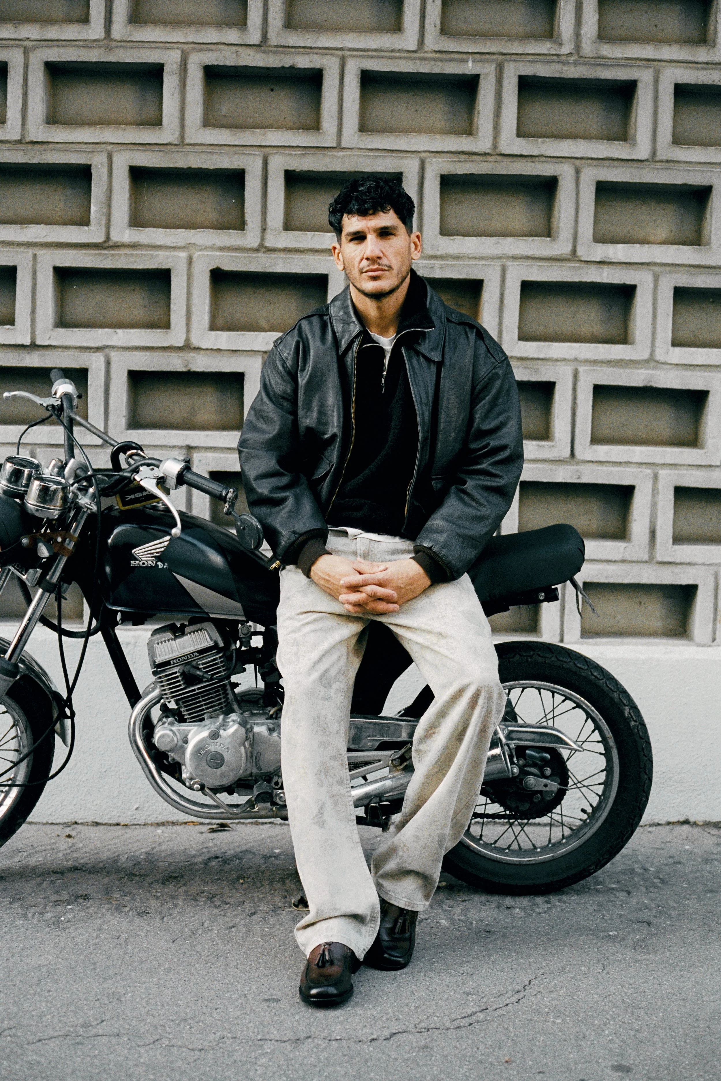 Un homme assis sur une moto noire, portant une veste en cuir noire et un pantalon beige, devant un mur en blocs de béton.