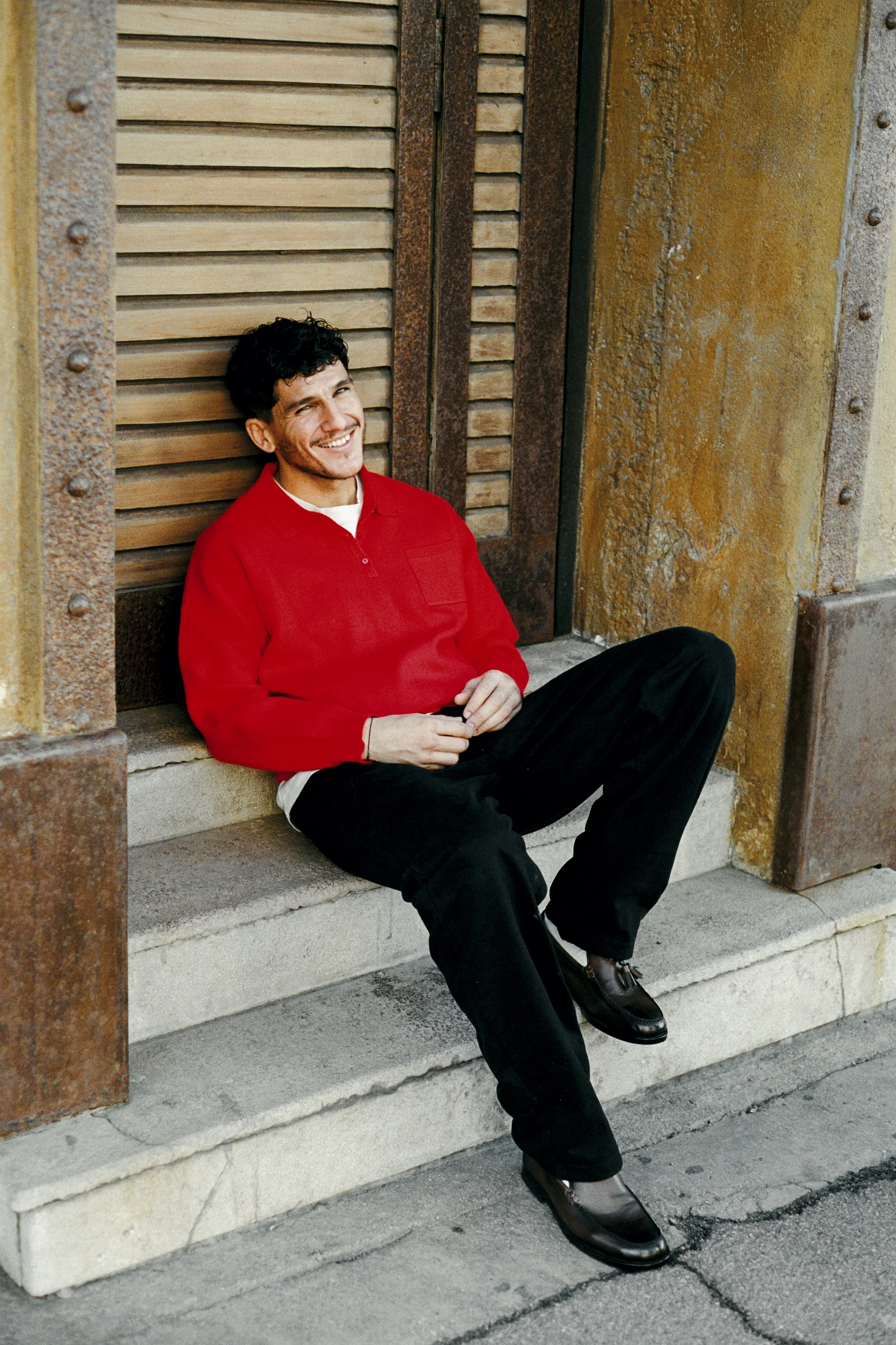 Un jeune homme souriant assis sur un escalier en pierre devant une façade en métal et en pierre. Il porte un pull rouge, un pantalon noir et des chaussures marron.
