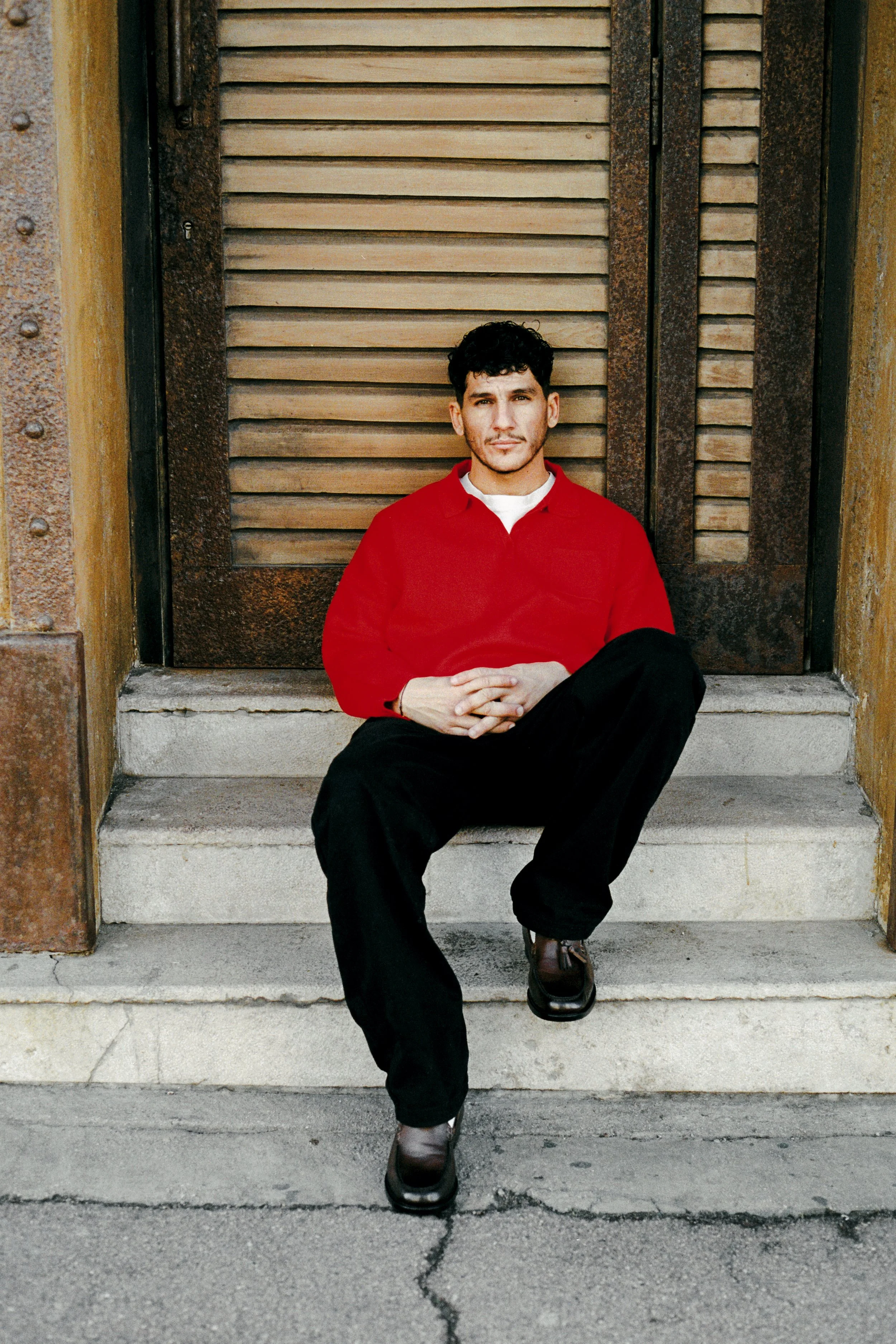 Un homme assis sur des marches en pierre devant une porte en bois, portant un pull rouge, un jean noir et des chaussures noires.