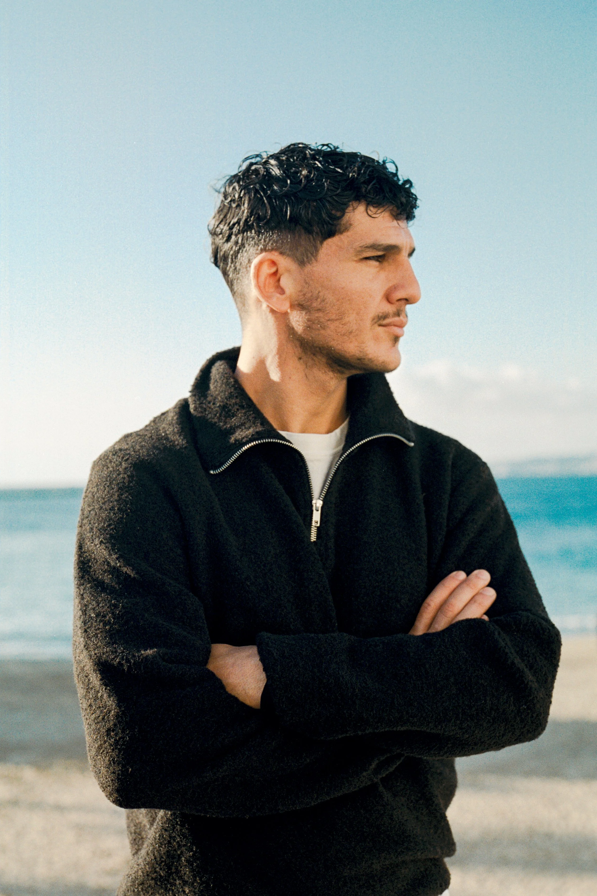 Un homme regarde vers la droite, les bras croisés, sur la plage avec la mer en arrière-plan, portant un pull noir avec un col zippé.