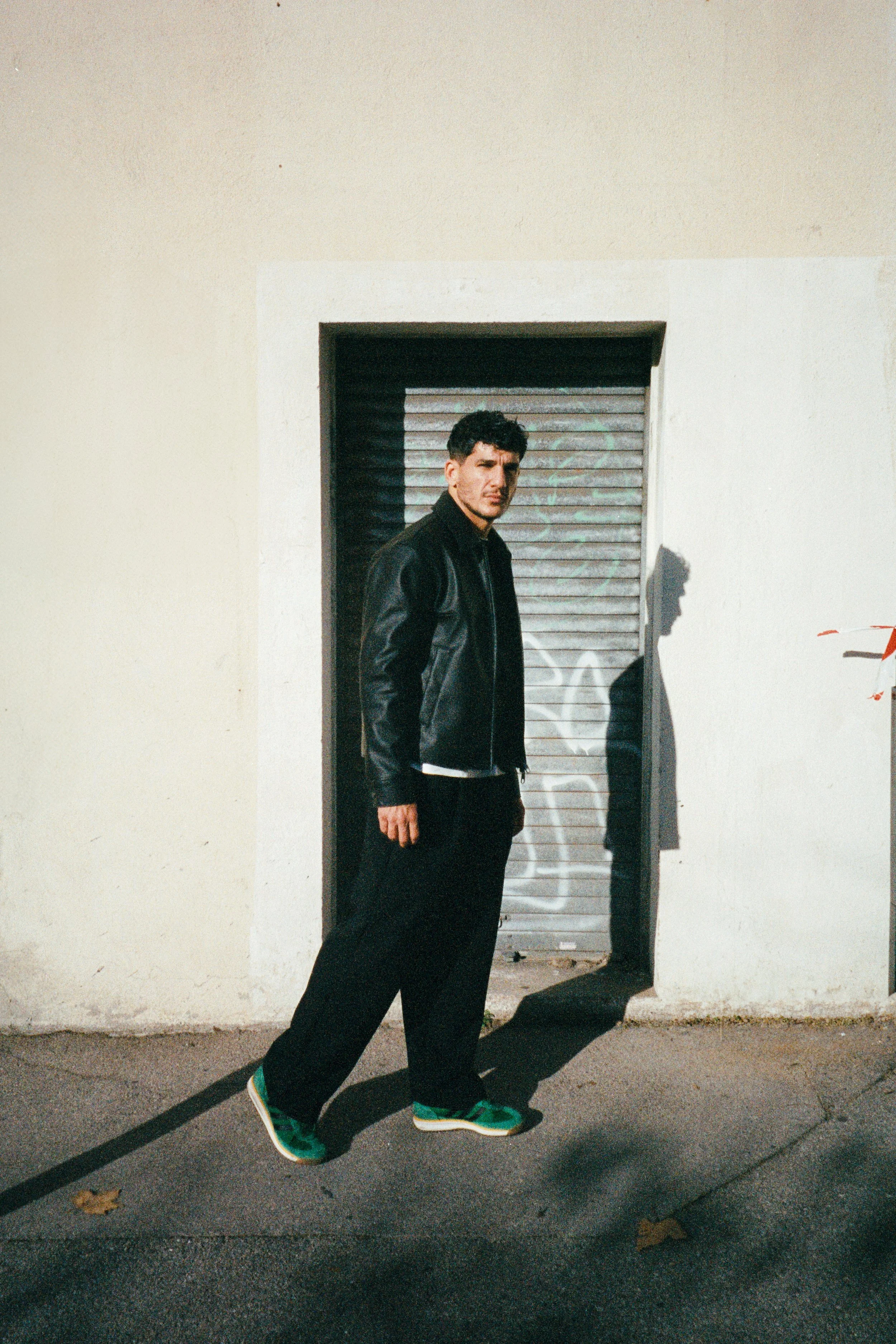 Un homme en veste en cuir noire, pantalon noir et chaussures vertes, se tient devant une porte métallique enroulée dans une rue urbaine, avec un mur beige à l'arrière-plan et la lumière du soleil créant une ombre visible.