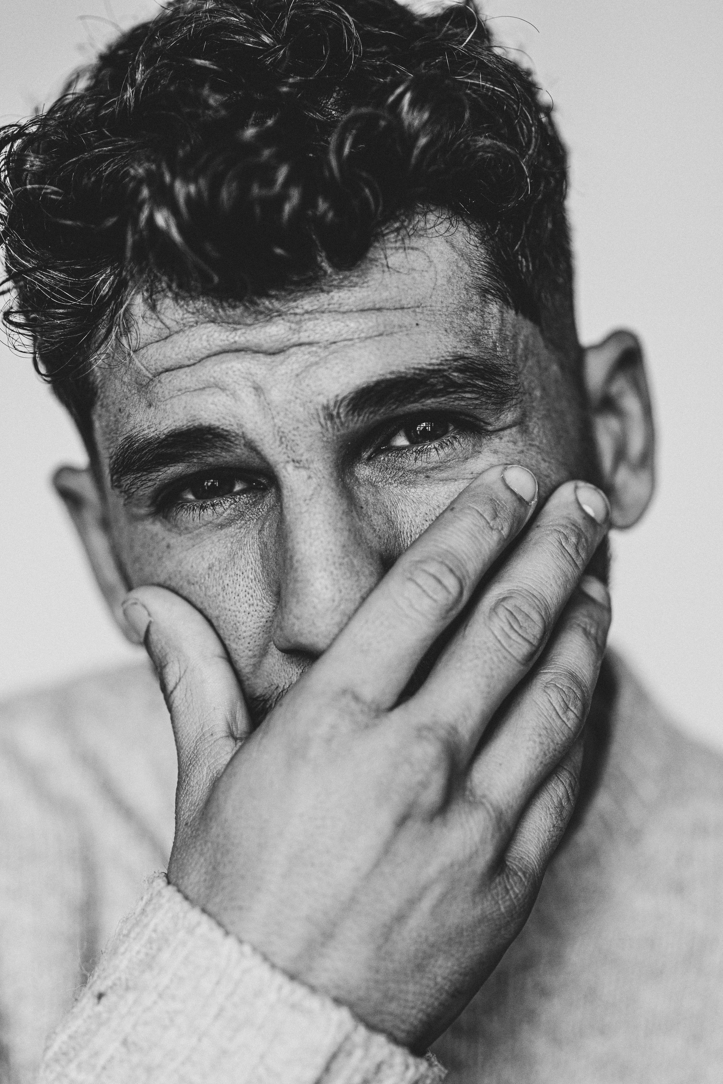 Photo en noir et blanc d'un homme avec des cheveux bouclés, tenant sa main contre son visage, regardant vers l'appareil.