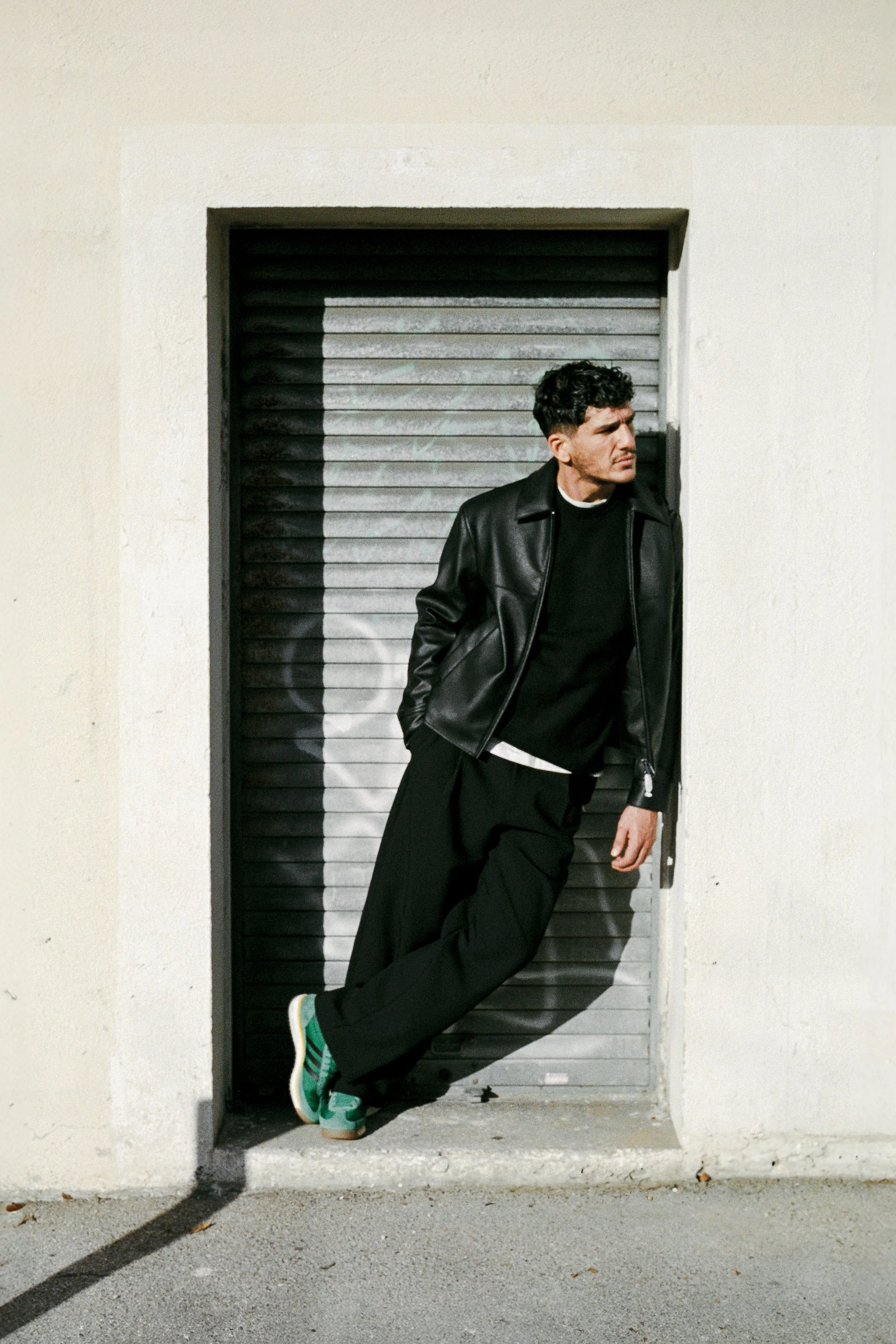 Un homme debout dans une entrée sombre, portant un blouson en cuir noir, un pull noir, un pantalon ample, et des chaussures de sport vertes. Il regarde vers la droite, avec un mur blanc derrière lui.