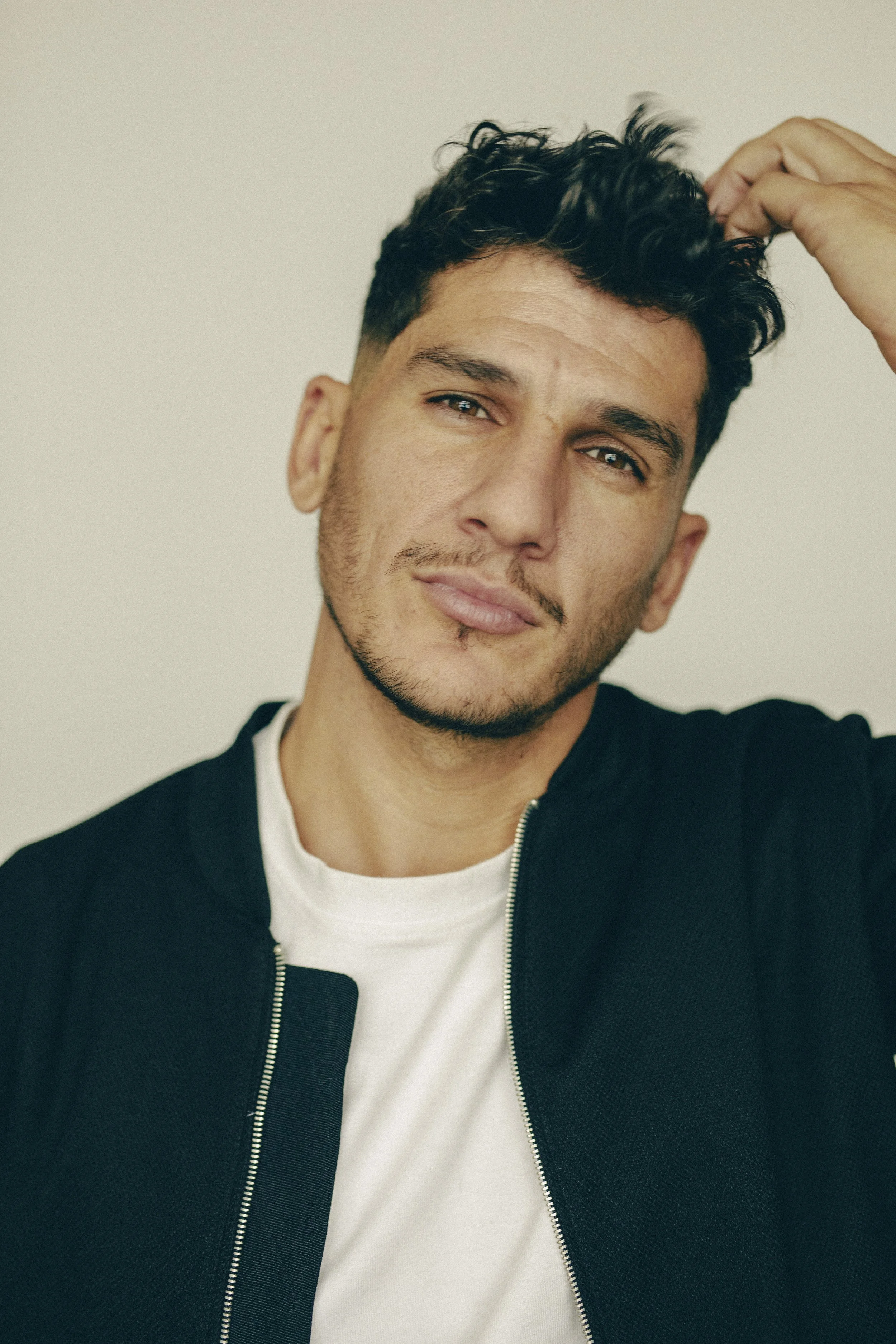 Jeune homme avec cheveux courts et barbe, portant un sweatshirt noir et un t-shirt blanc, touche ses cheveux, regarde vers l'appareil photo.