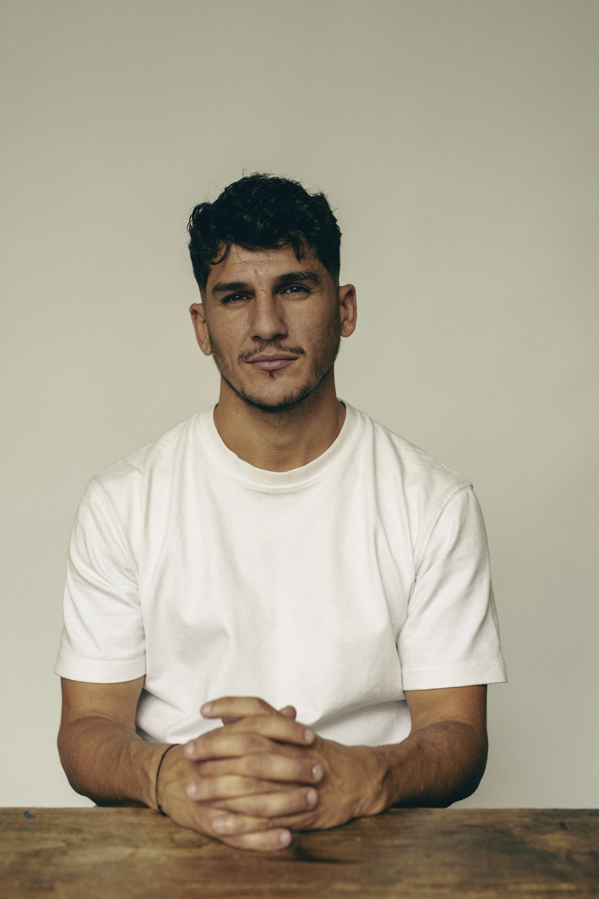Un homme assis à une table en bois, portant un t-shirt blanc, avec des cheveux courts et bouclés, regarde l'appareil photo, souriant légèrement, dans un fond neutre beige.