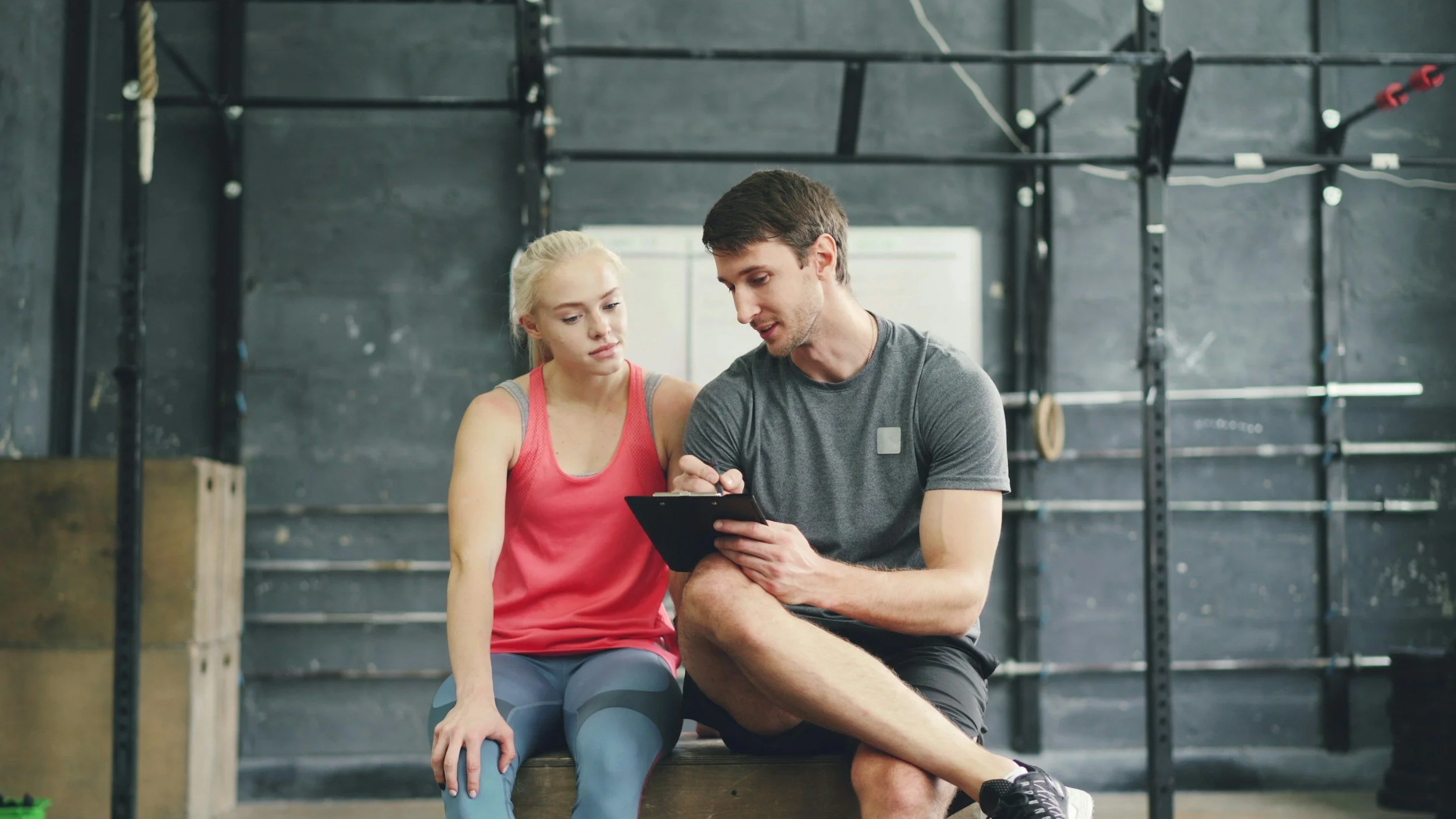Een man en een vrouw in een sportschool die samen naar een tablet kijken, de vrouw is in sportkleding en de man houdt het apparaat vast.