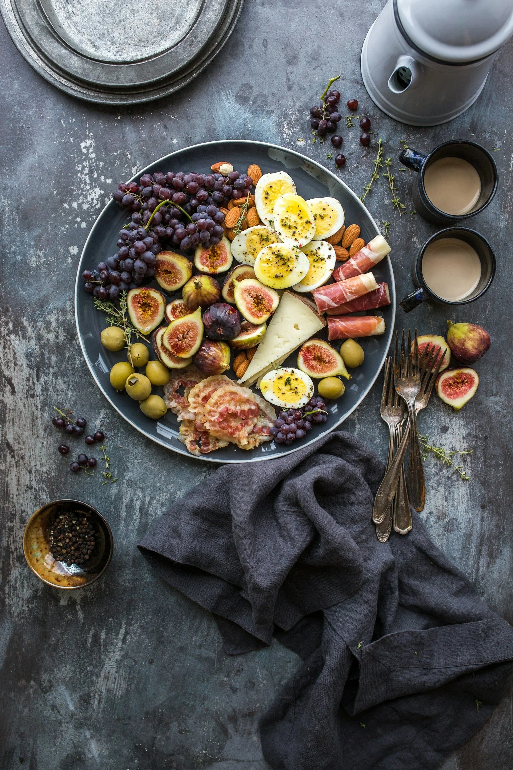 Een bord met verschillende soorten fruit zoals vijgen, druiven, pruimen en een gekookt ei, geserveerd met stukjes kaas en vleeswaren, op een donkere tafel met bestek, kopjes koffie of thee en een zwarte doek.