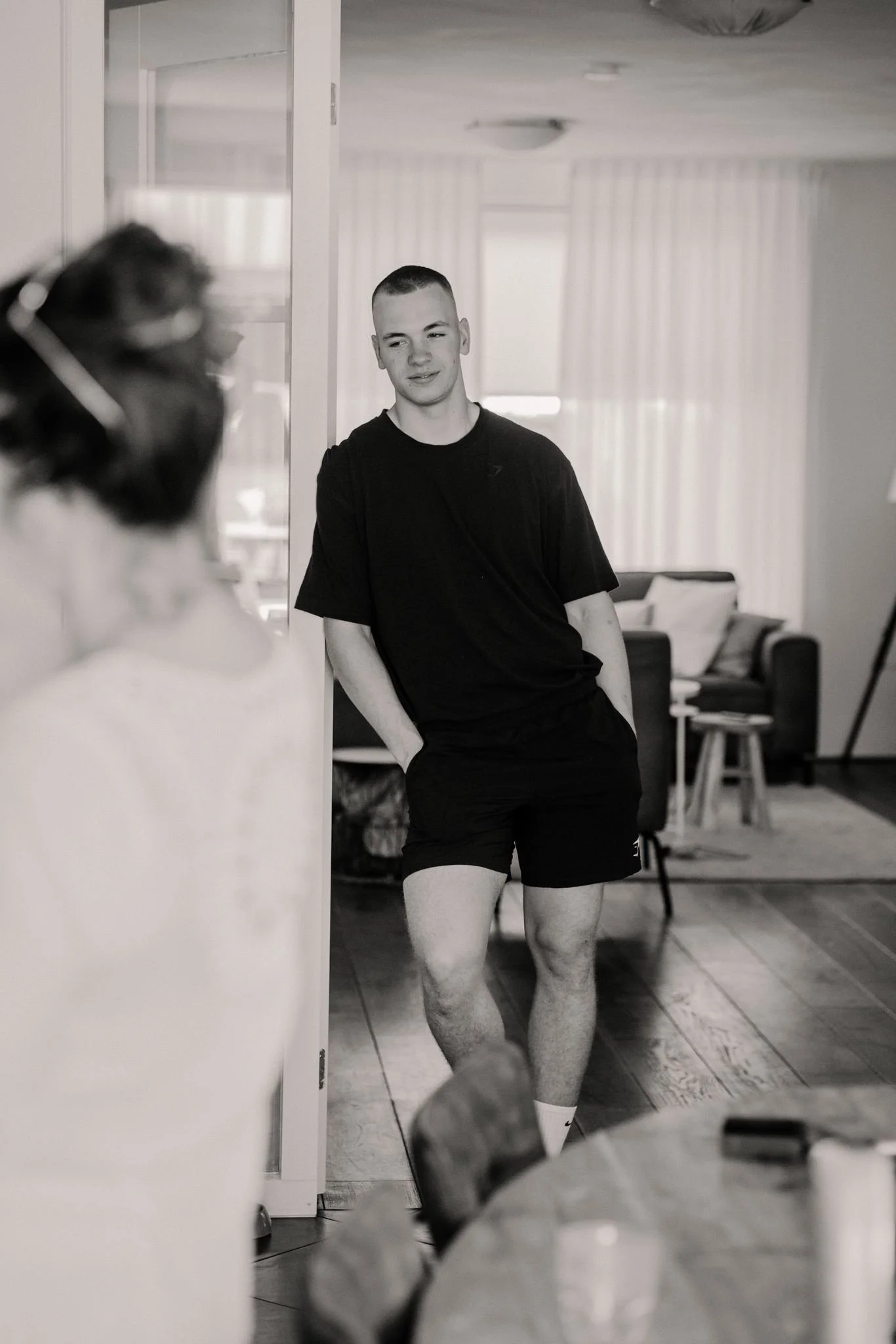 Een jonge man staat binnen in een kamer, leunend tegen de muur, met zijn handen in de zakken van zijn korte broek, terwijl hij naar een persoon kijkt die op de voorgrond niet volledig zichtbaar is.