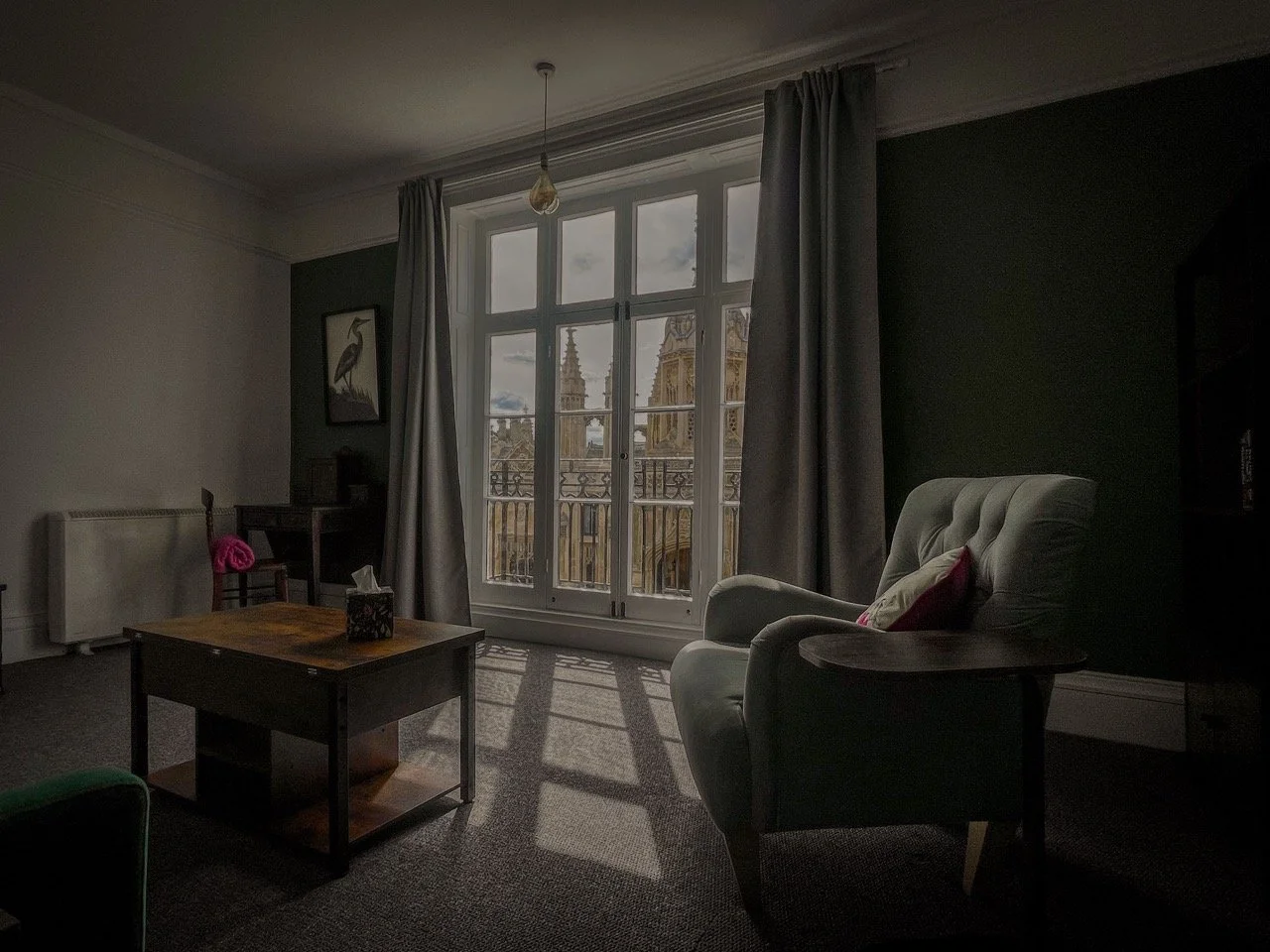 A living room with a large window view of cathedral spires, a cozy armchair with a pillow, a coffee table, curtains, and a small desk with a framed bird picture on the wall.