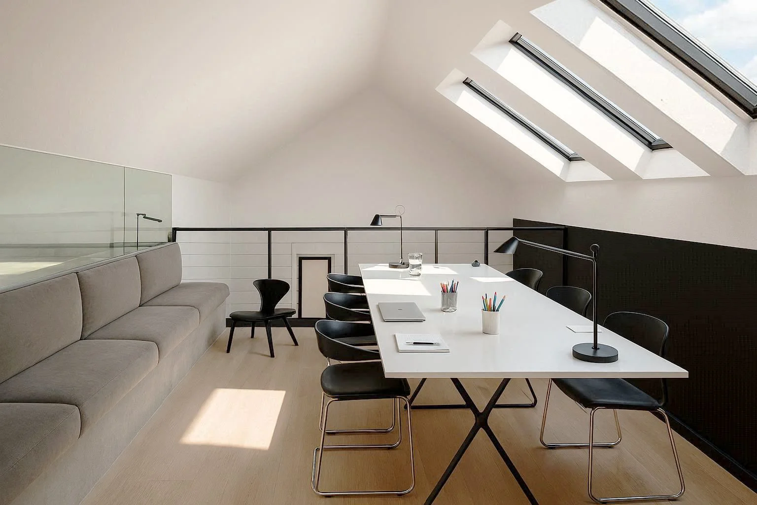Salle de réunion ou bureau avec une grande table blanche, plusieurs chaises noires, un canapé gris sur le côté, deux lampes de bureau noires, stylos de différentes couleurs dans un pot, un ordinateur portable, un verre d'eau, sous un plafond en pente avec deux fenêtres de toit laissant entrer la lumière naturelle.