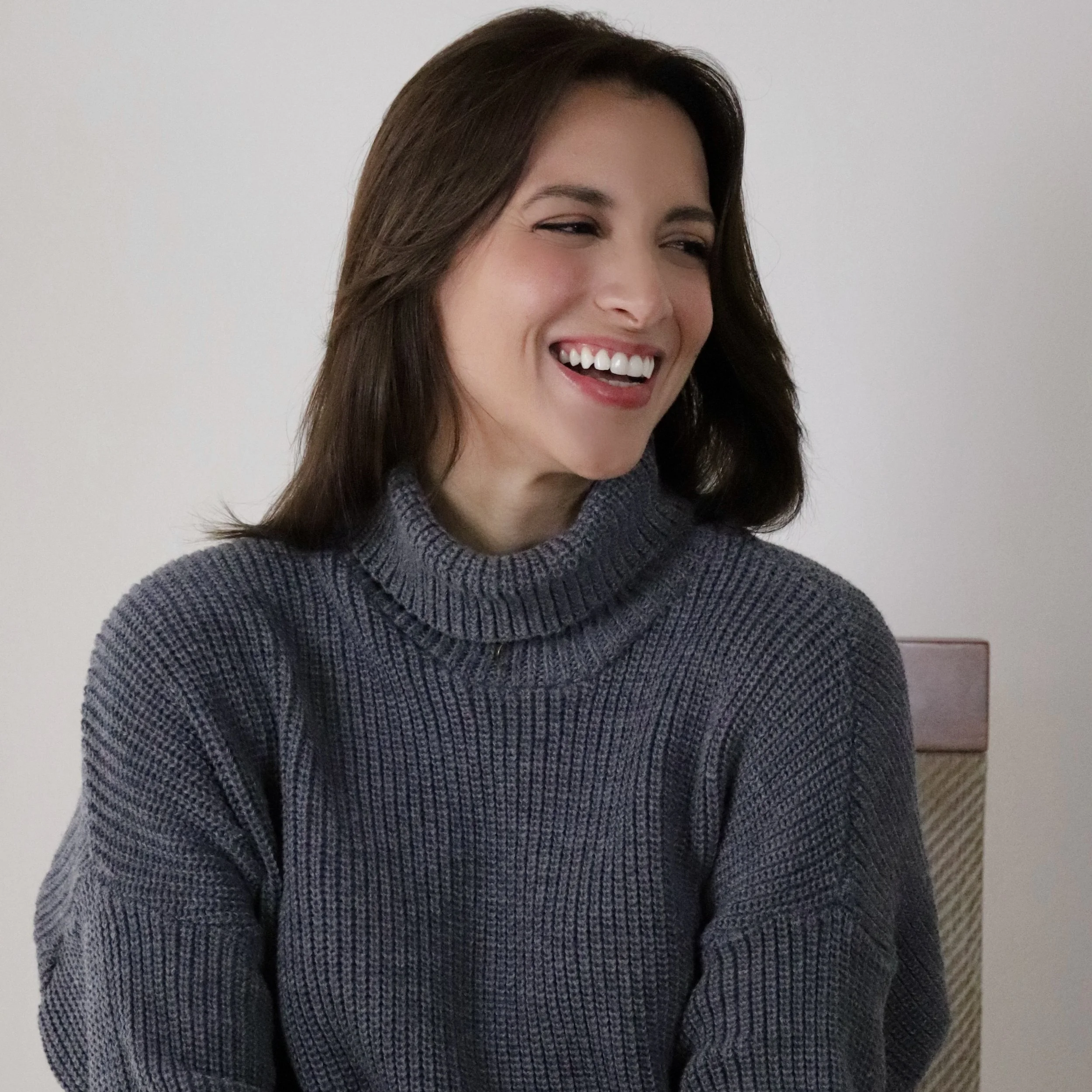 A woman with shoulder-length brown hair, smiling and wearing a gray turtleneck sweater, sitting on a chair against a white background.