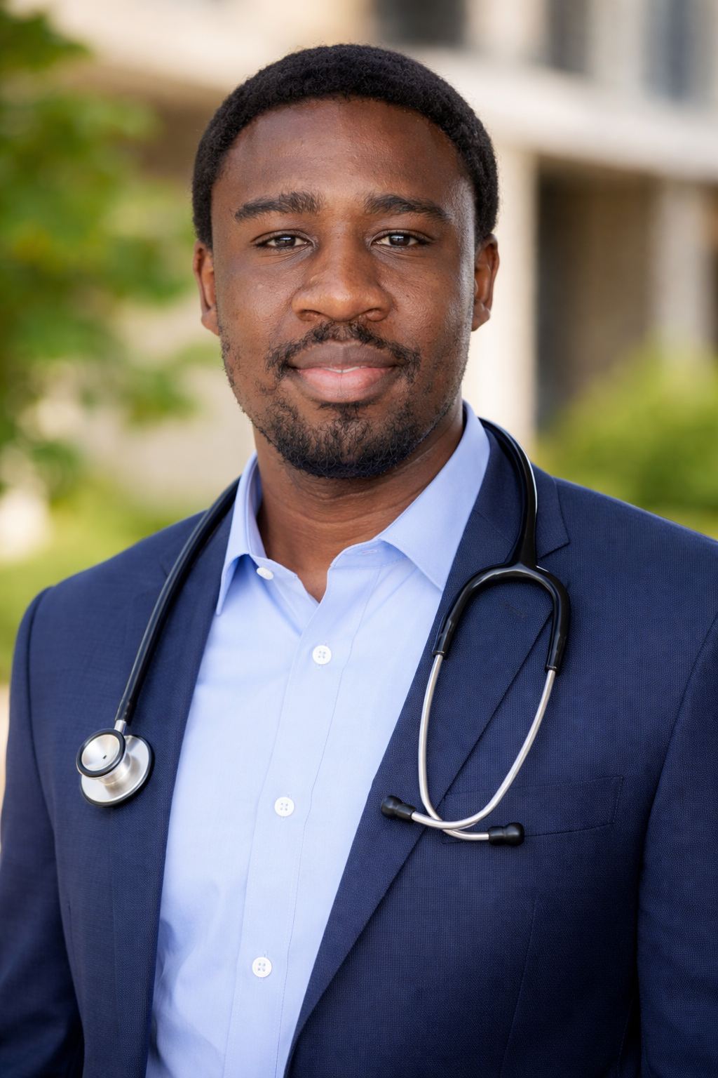 A male doctor or healthcare professional outdoors, wearing a blue blazer and a light blue shirt, with a stethoscope around his neck, smiling and looking at the camera.