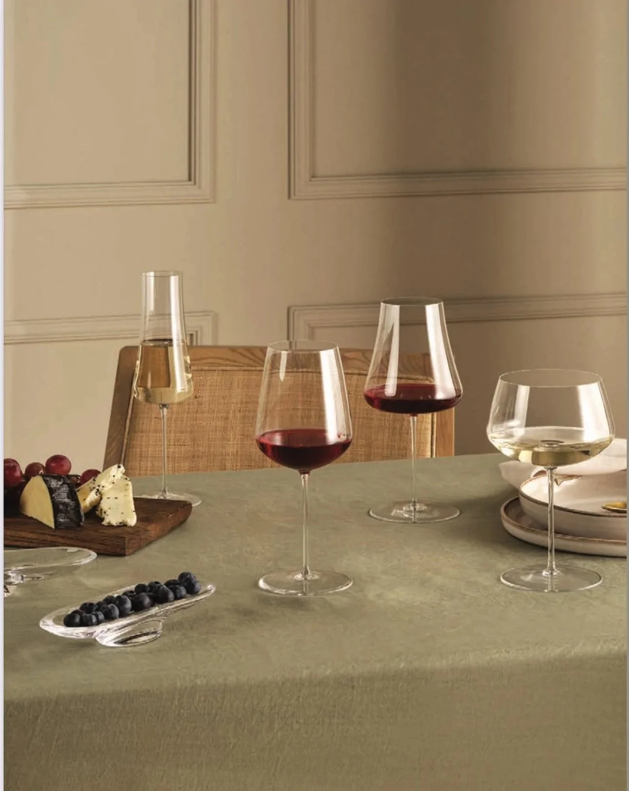 A table set with four wine glasses containing white and red wine, a small plate with blueberries, a cheese platter with grapes and cheese, and a white plate with a gold spoon, on a green tablecloth.