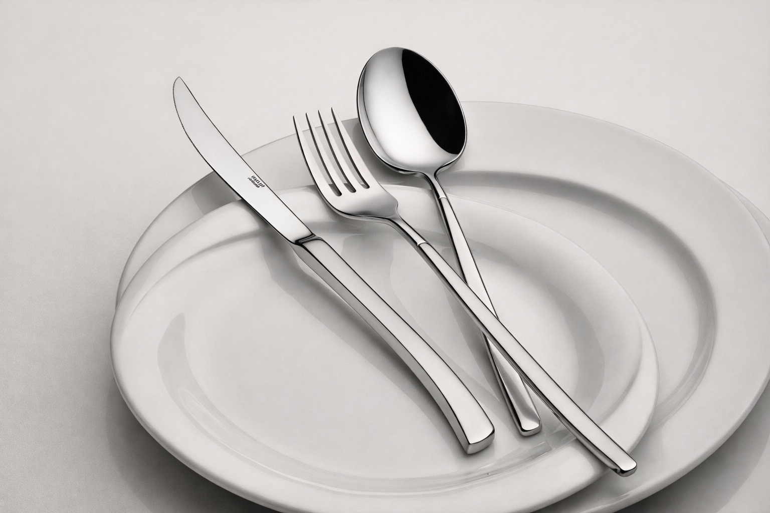 A white ceramic plate with a matching smaller plate on top, topped with a stainless steel knife, fork, and spoon on a light-colored surface.