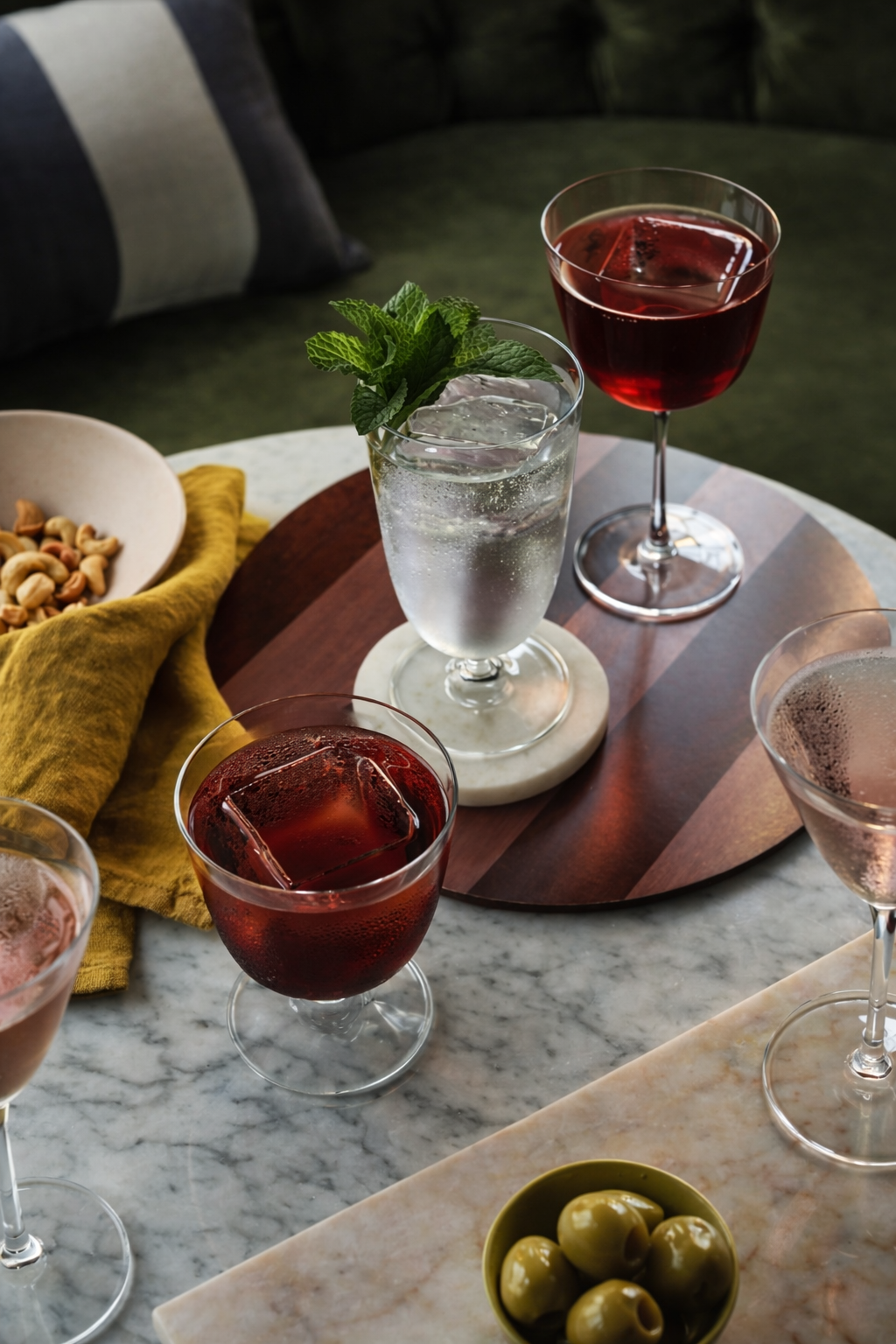 Set of drinks including two glasses of red wine and a glass of sparkling water with a mint garnish on a marble table, along with a bowl of mixed nuts, a bowl of green olives, and a dish with a pink cocktail.