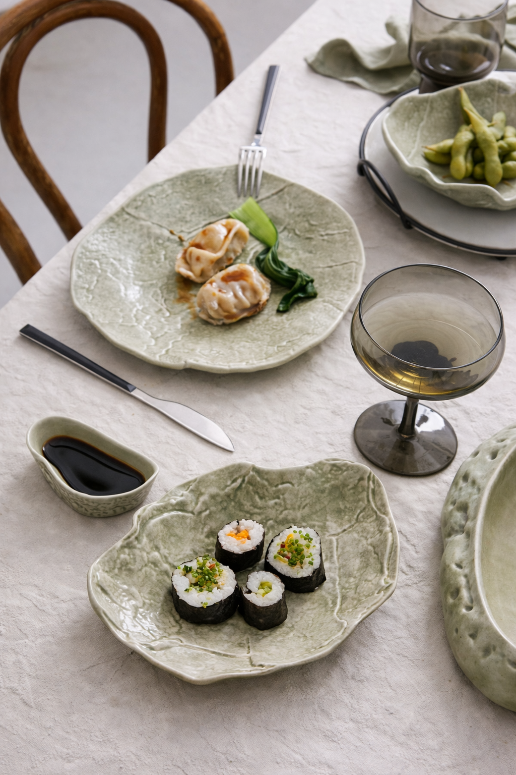 A top view of a dining table with sushi rolls, dumplings, soy sauce, and a glass of white wine on a beige tablecloth.