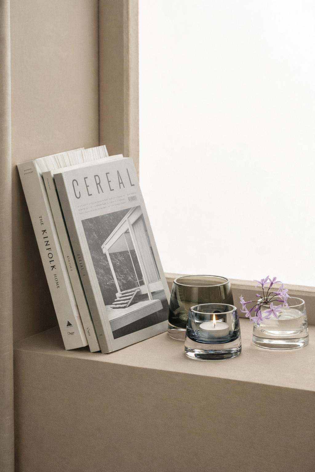 A windowsill with books, candles, and a small glass vase with purple flowers in water.